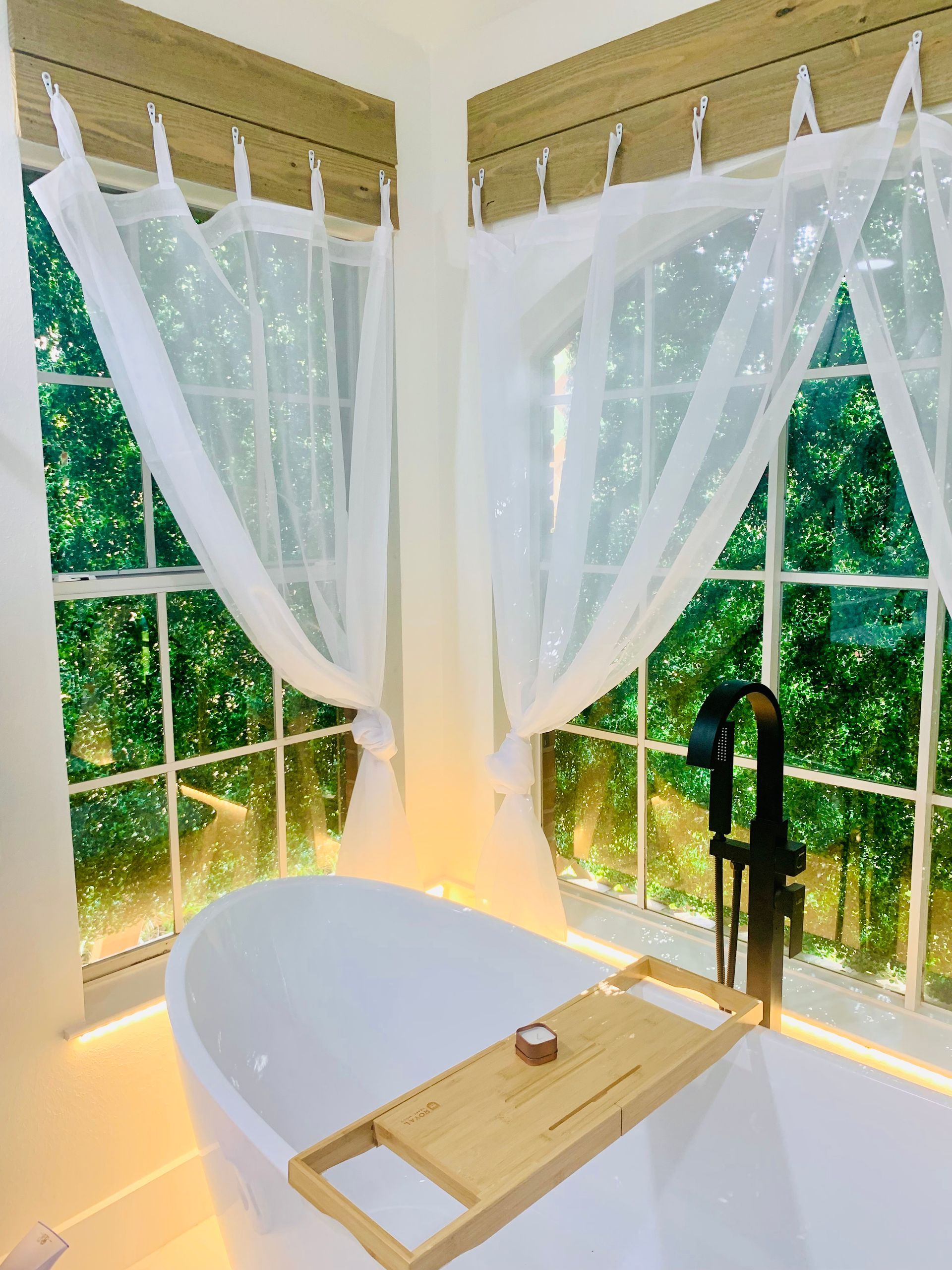 A white bathtub with a wooden tray sits beneath two windows with sheer curtains, overlooking greenery.