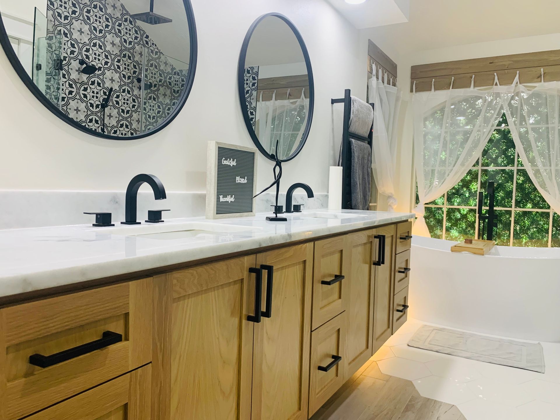 Bathroom with wooden vanity, black fixtures, round mirrors, and a window with sheer curtains.