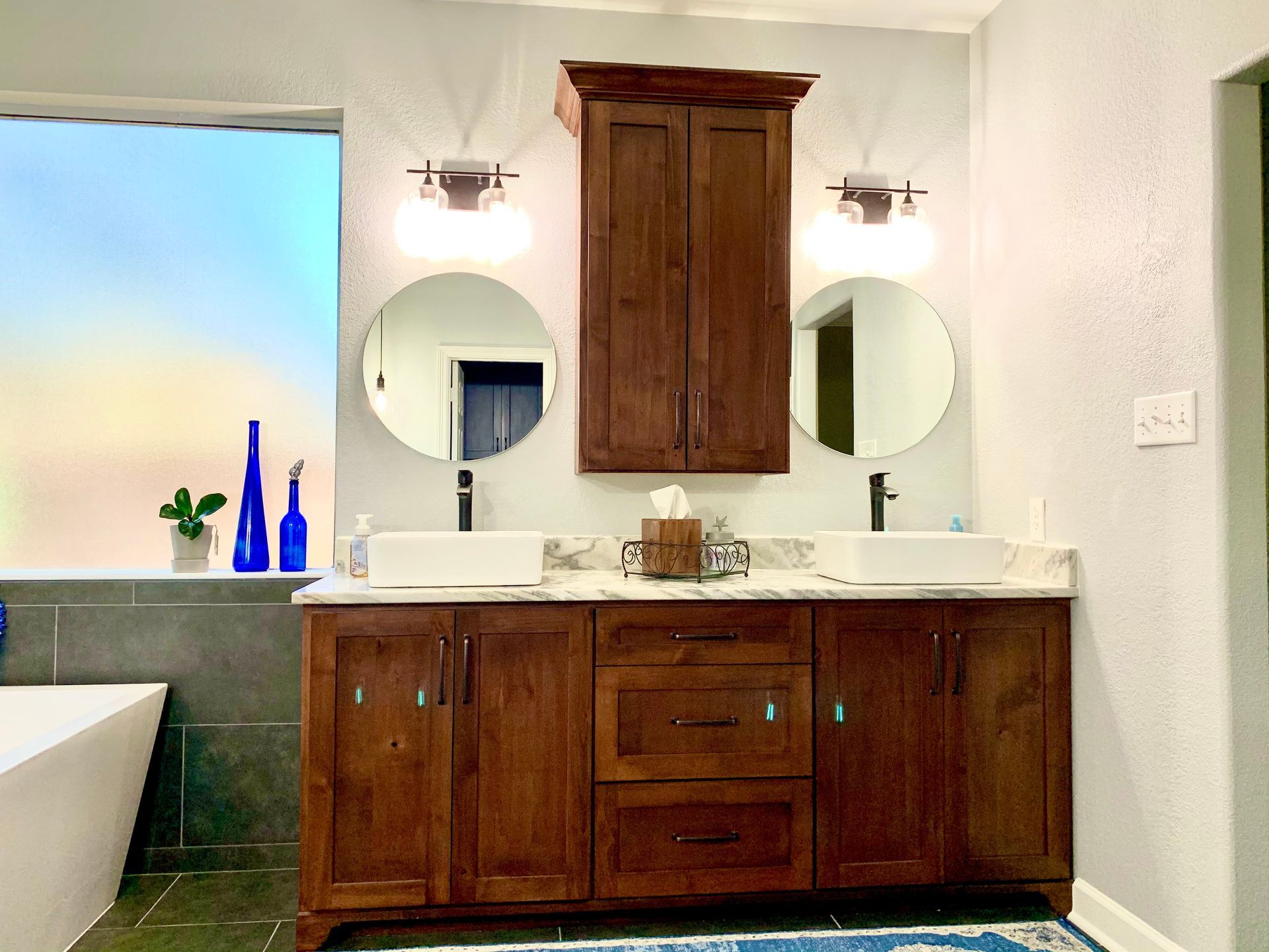 Bathroom with brown vanity, round mirrors, and a frosted window.