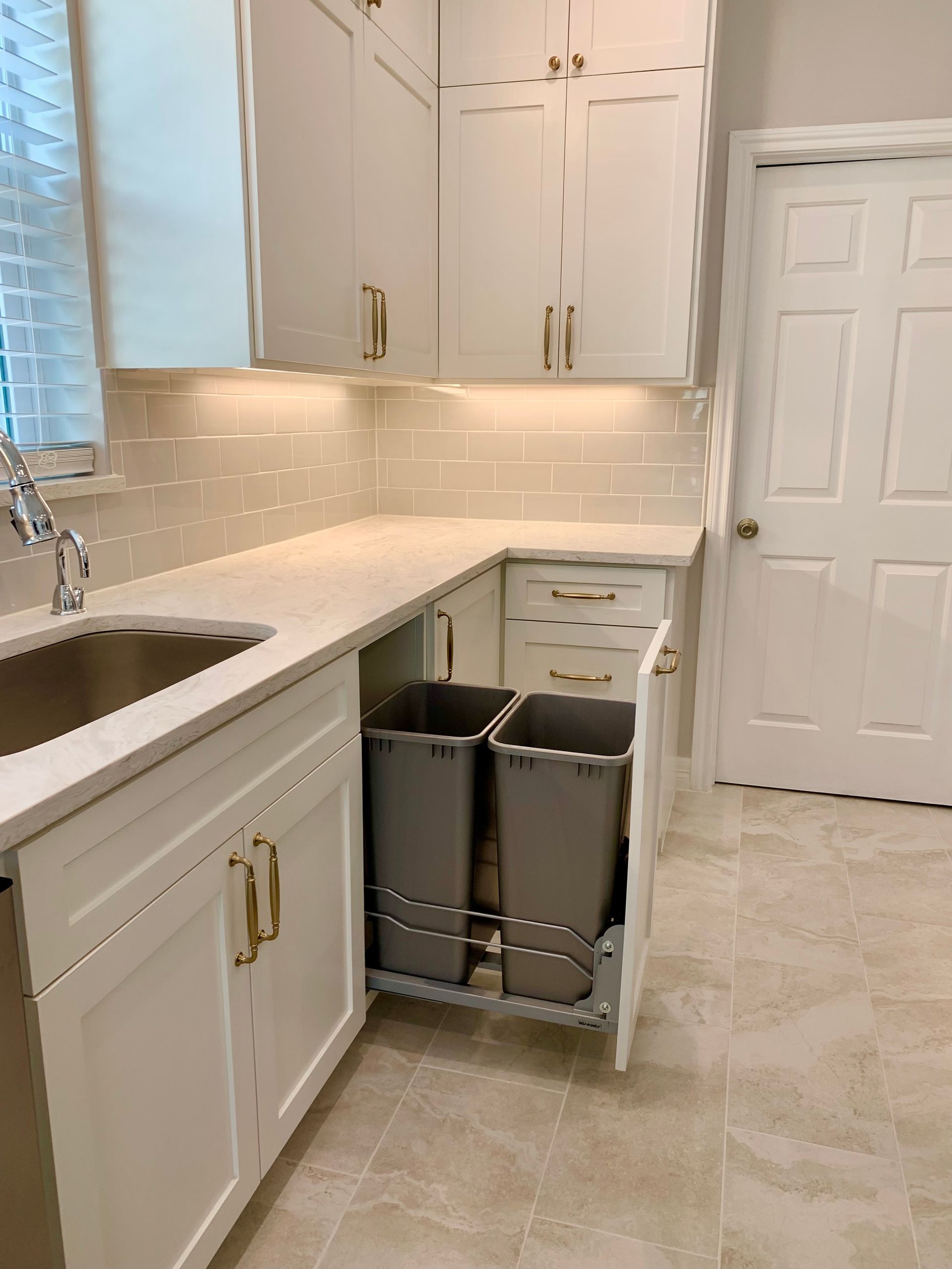 White kitchen cabinets with pull-out trash cans. Countertop and beige tile floor.