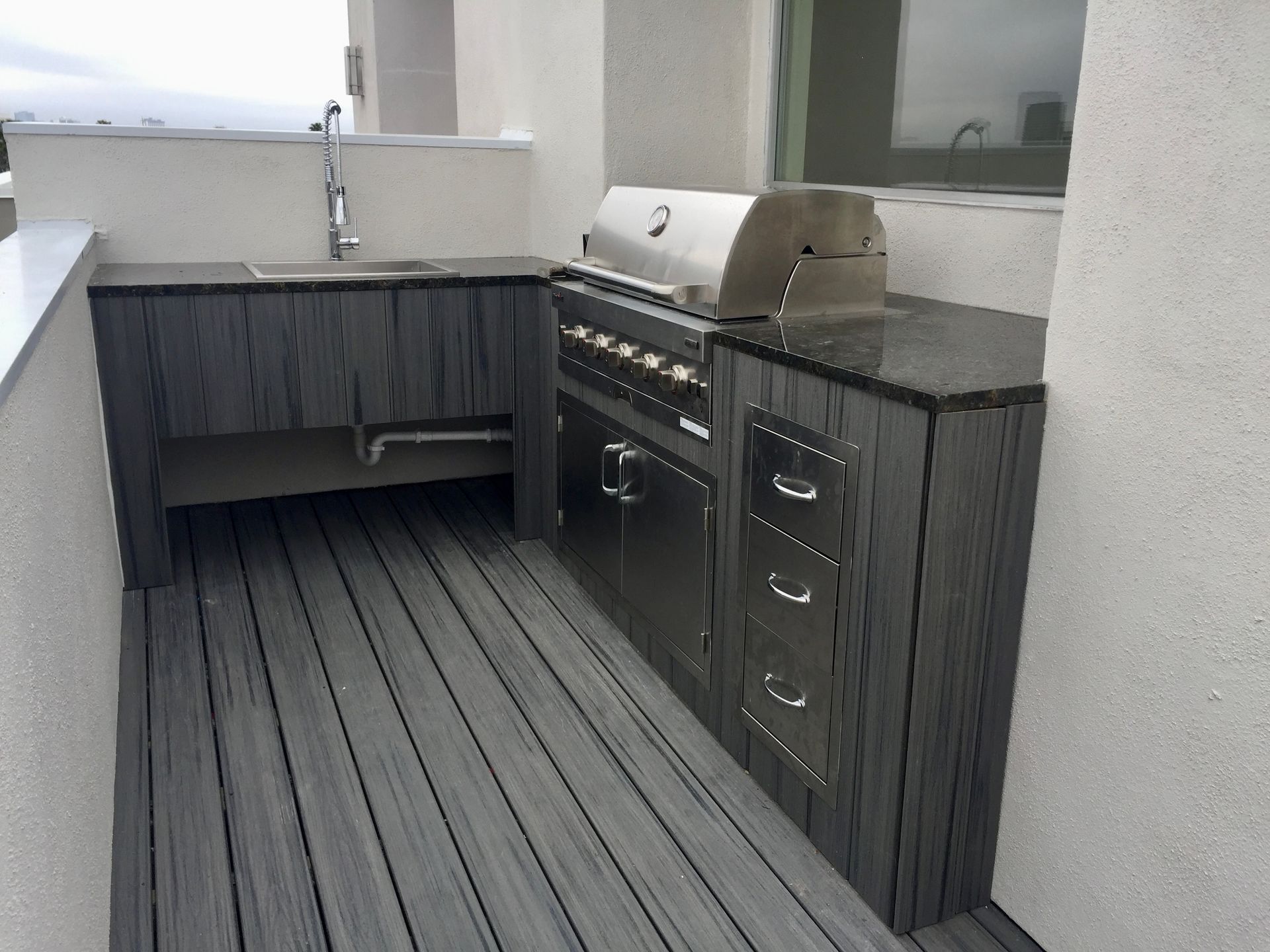 Outdoor kitchen with a stainless steel grill, cabinets, and a sink, on a gray deck.