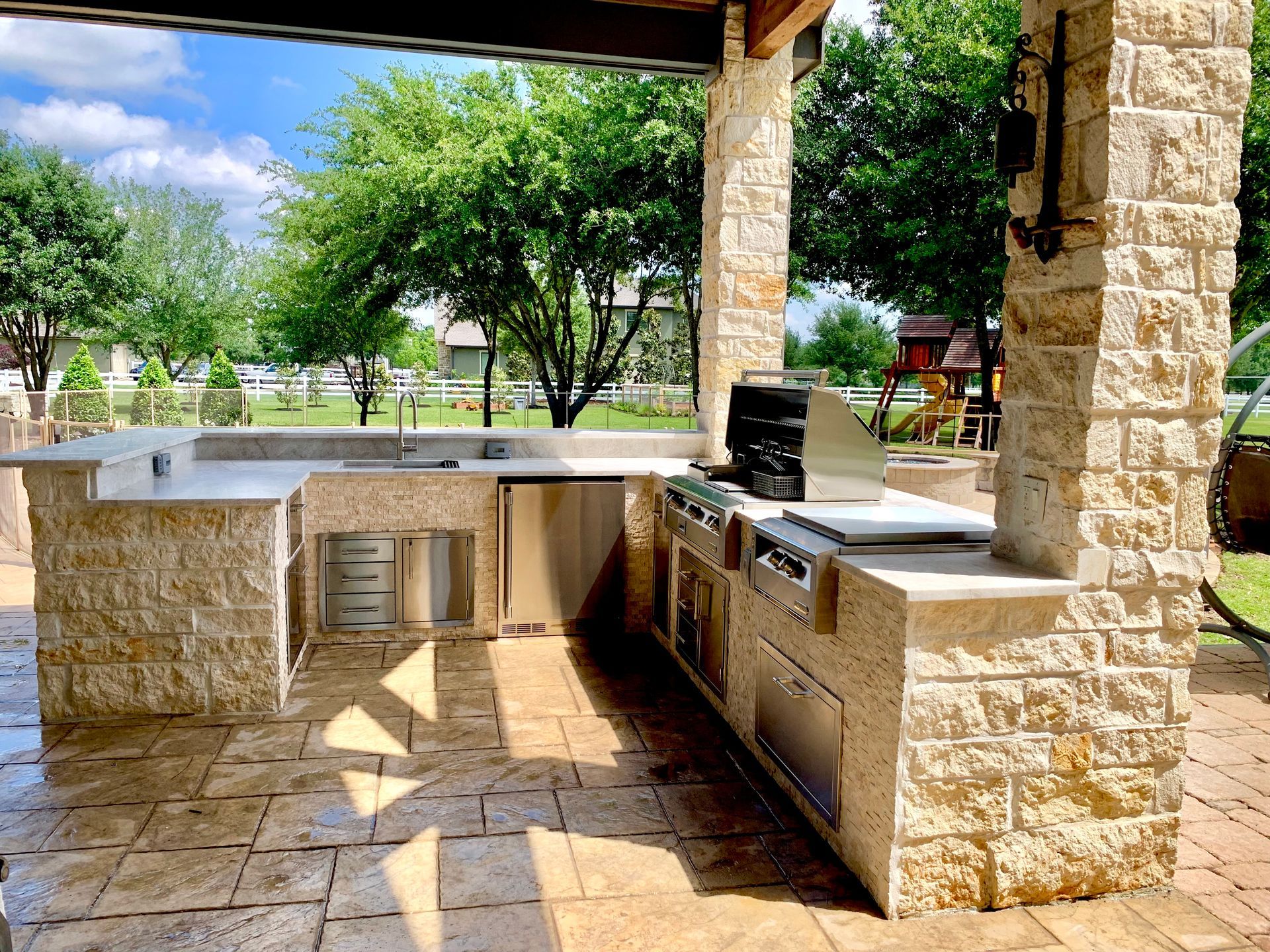 Outdoor kitchen with a stone countertop, grill, and sink, in a patio setting with trees in the background.