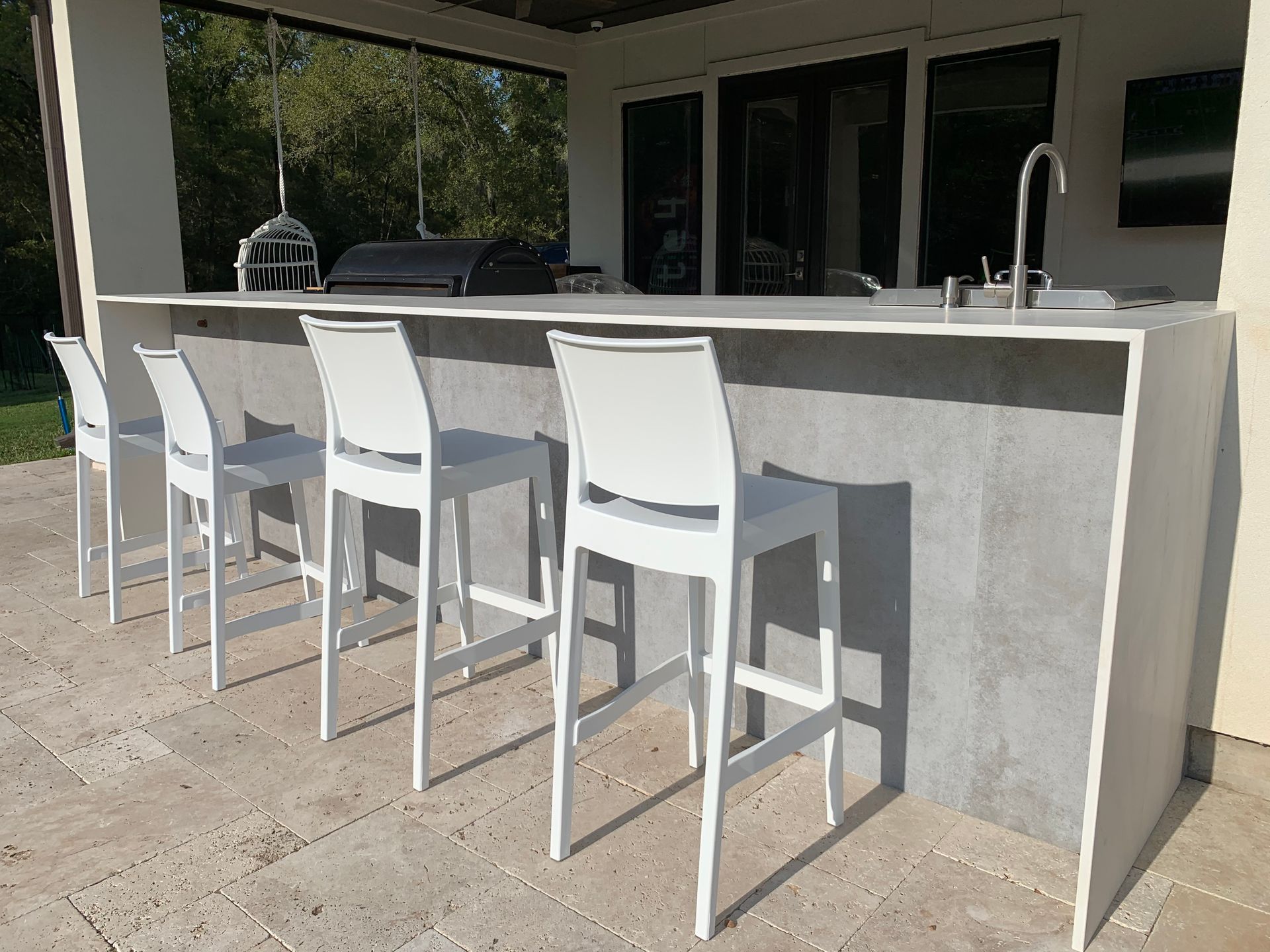 Outdoor kitchen with white bar stools, sink, and grill under a covered patio.