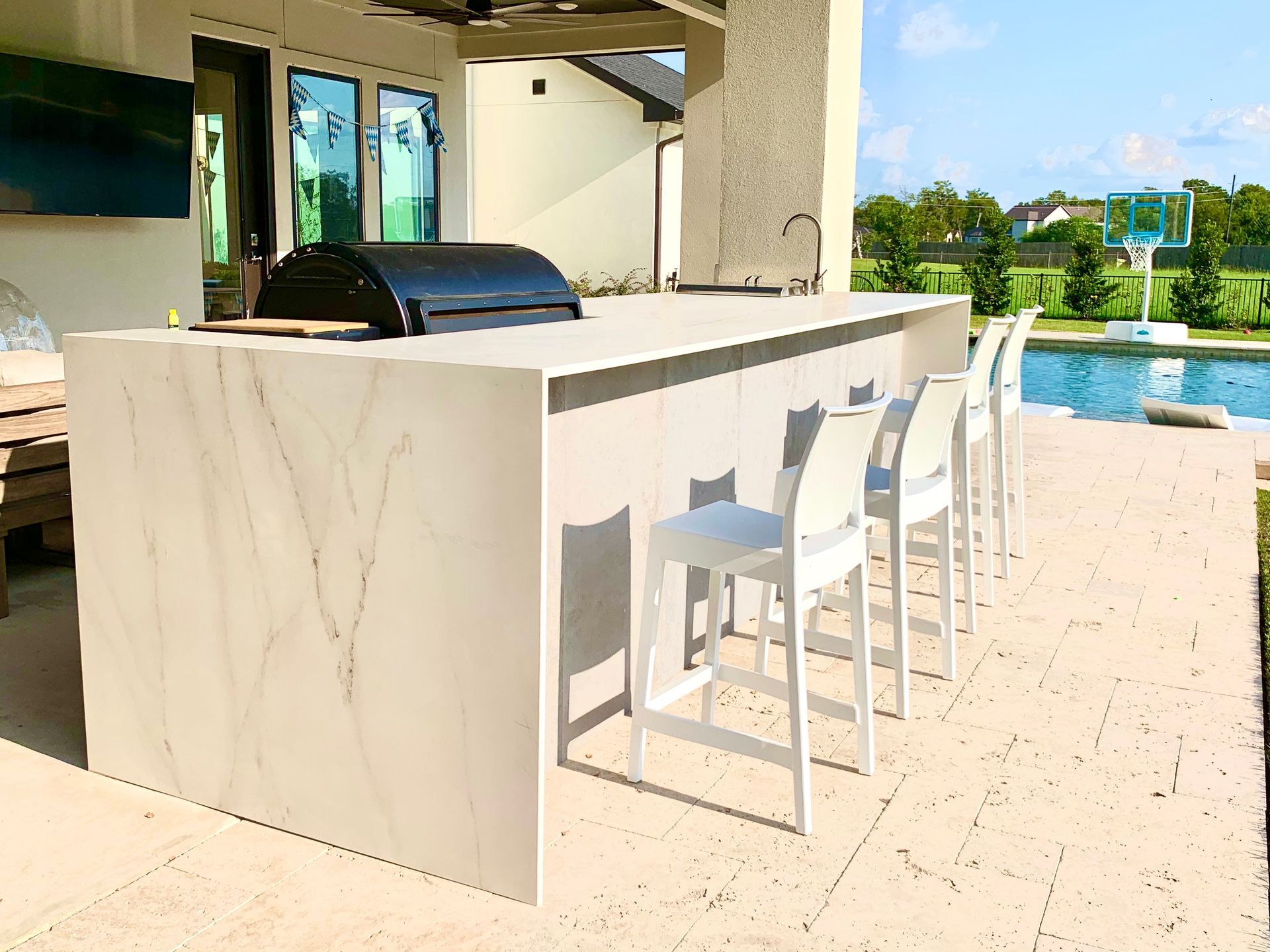 Outdoor kitchen with a grill, bar, white chairs, and a pool in the background.