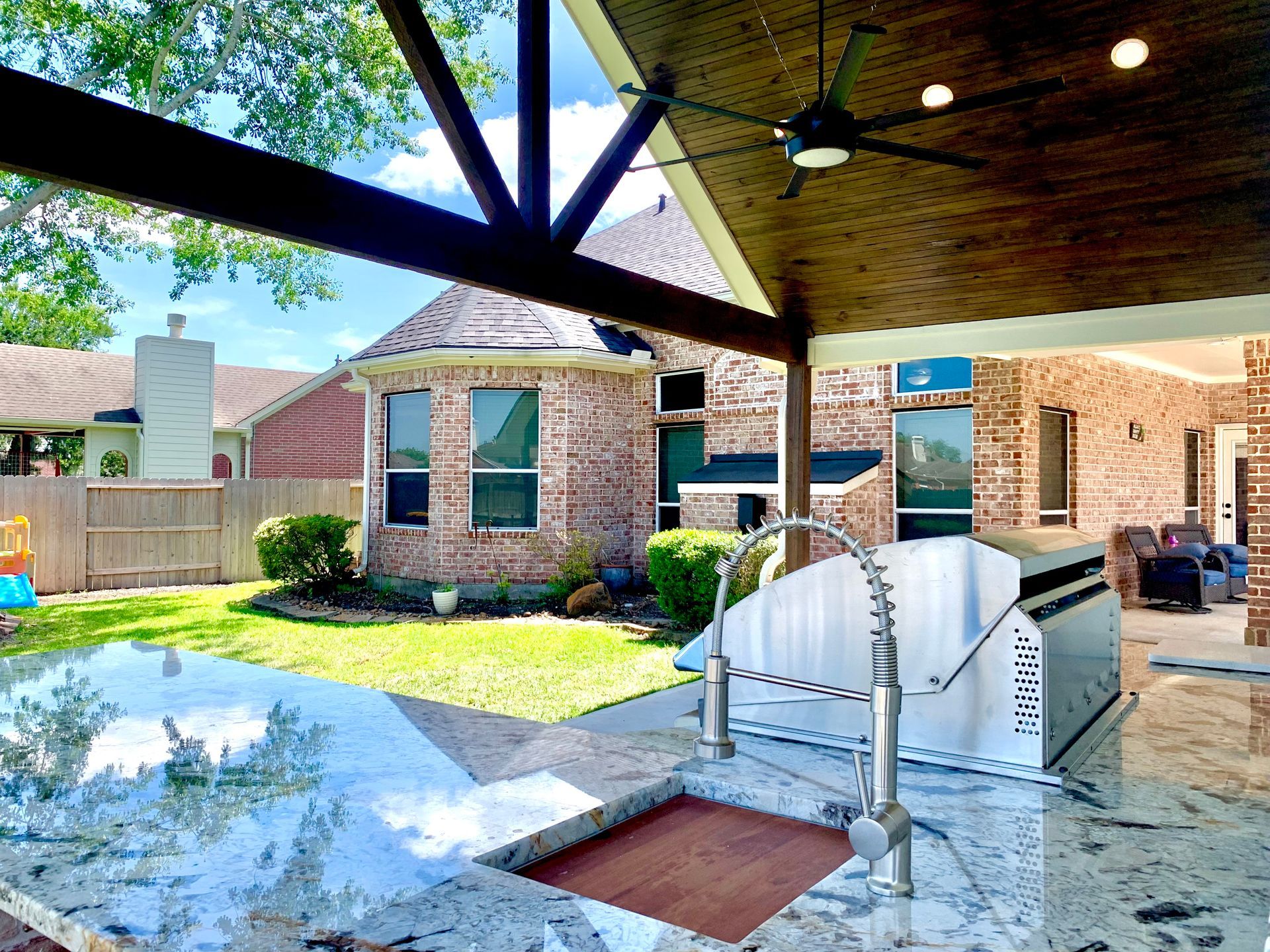 Outdoor kitchen under a wooden pergola, overlooking a brick house and backyard with green grass.