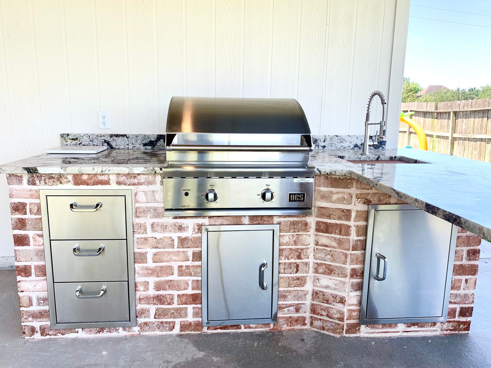 Outdoor kitchen with brick facade, stainless steel grill, drawers, and sink with granite countertops.