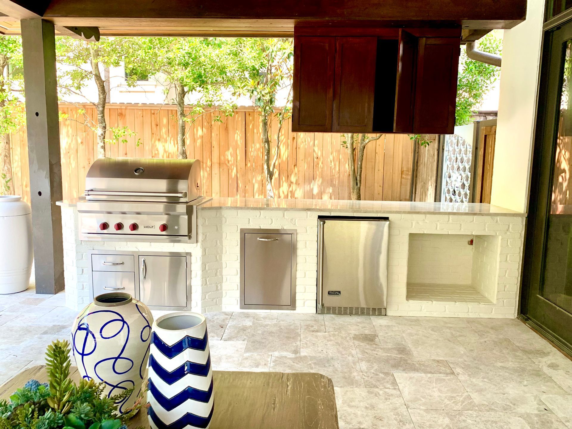 Outdoor kitchen with a grill, cabinets, and a refrigerator, set against a wooden fence.