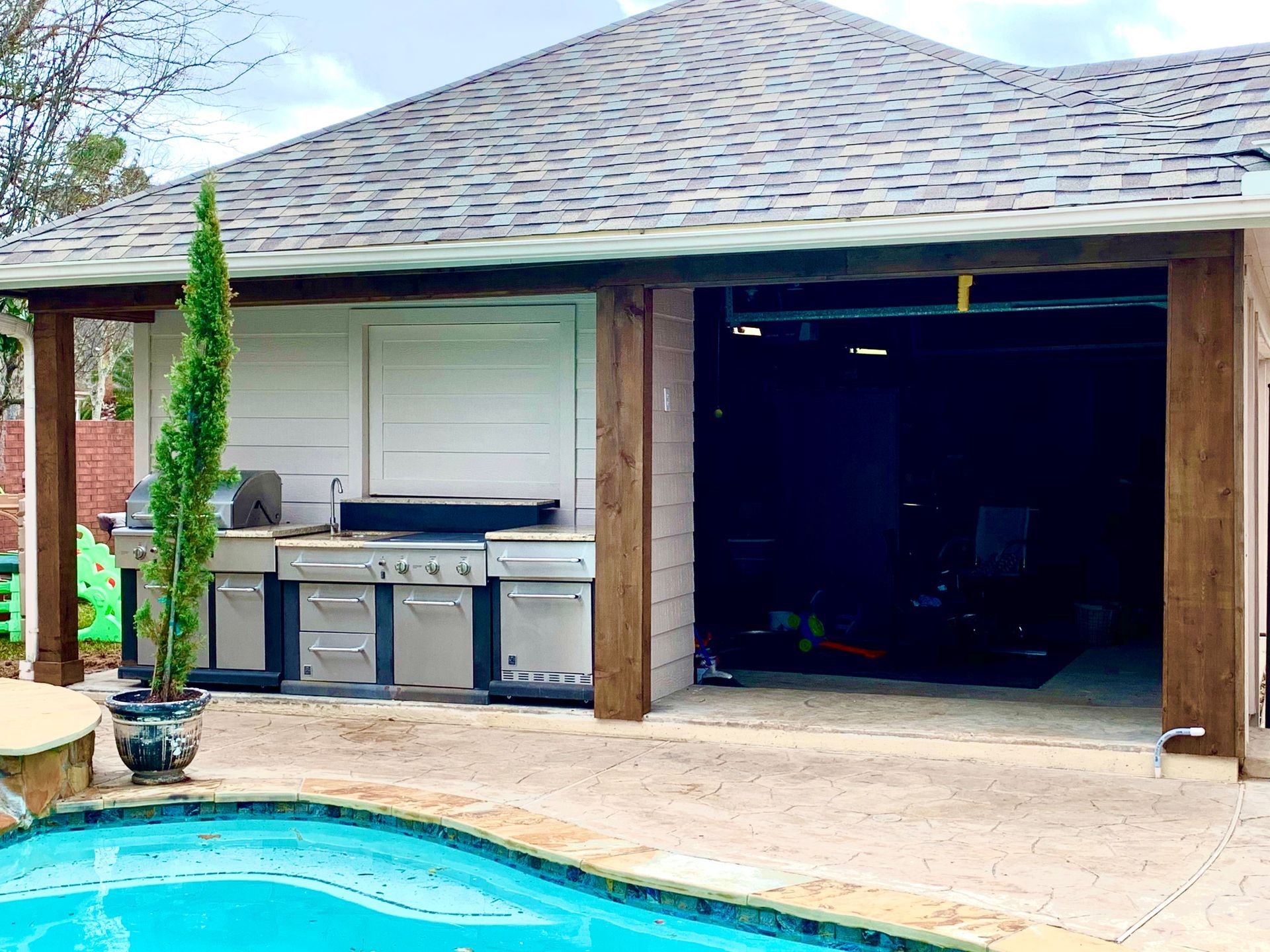 Outdoor kitchen with stainless steel grill, adjacent to a pool and a garage-like structure.