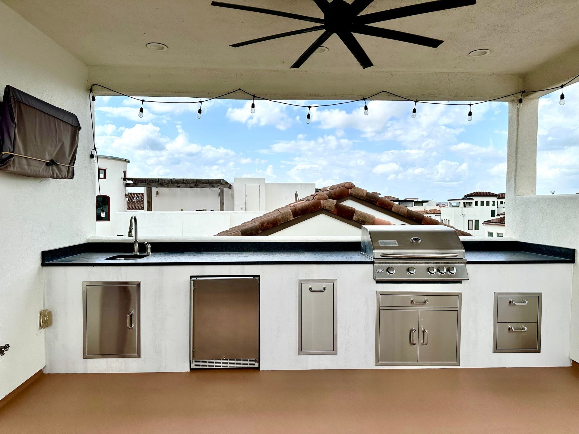 Outdoor kitchen with grill, sink, and storage against a white wall; brown floor, blue sky.