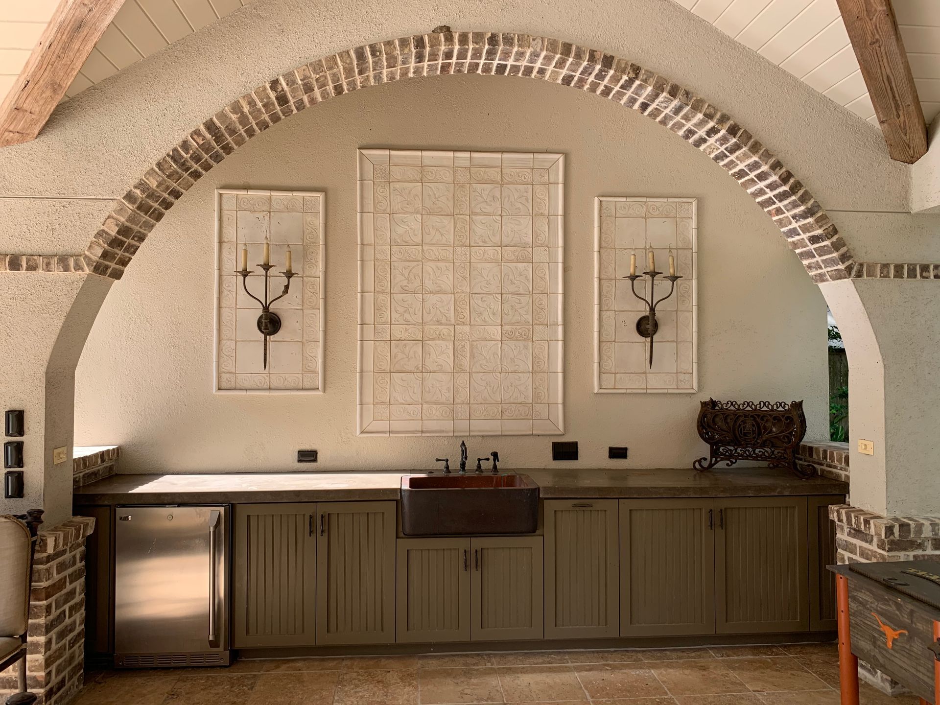 Outdoor kitchen with arched brick detail, olive cabinets, stainless steel appliances, and decorative wall art.