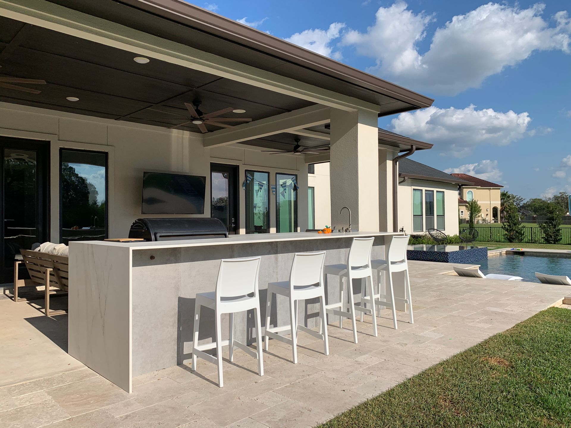 Outdoor kitchen with a bar, chairs, grill, TV, and pool. Sunny day with blue sky.
