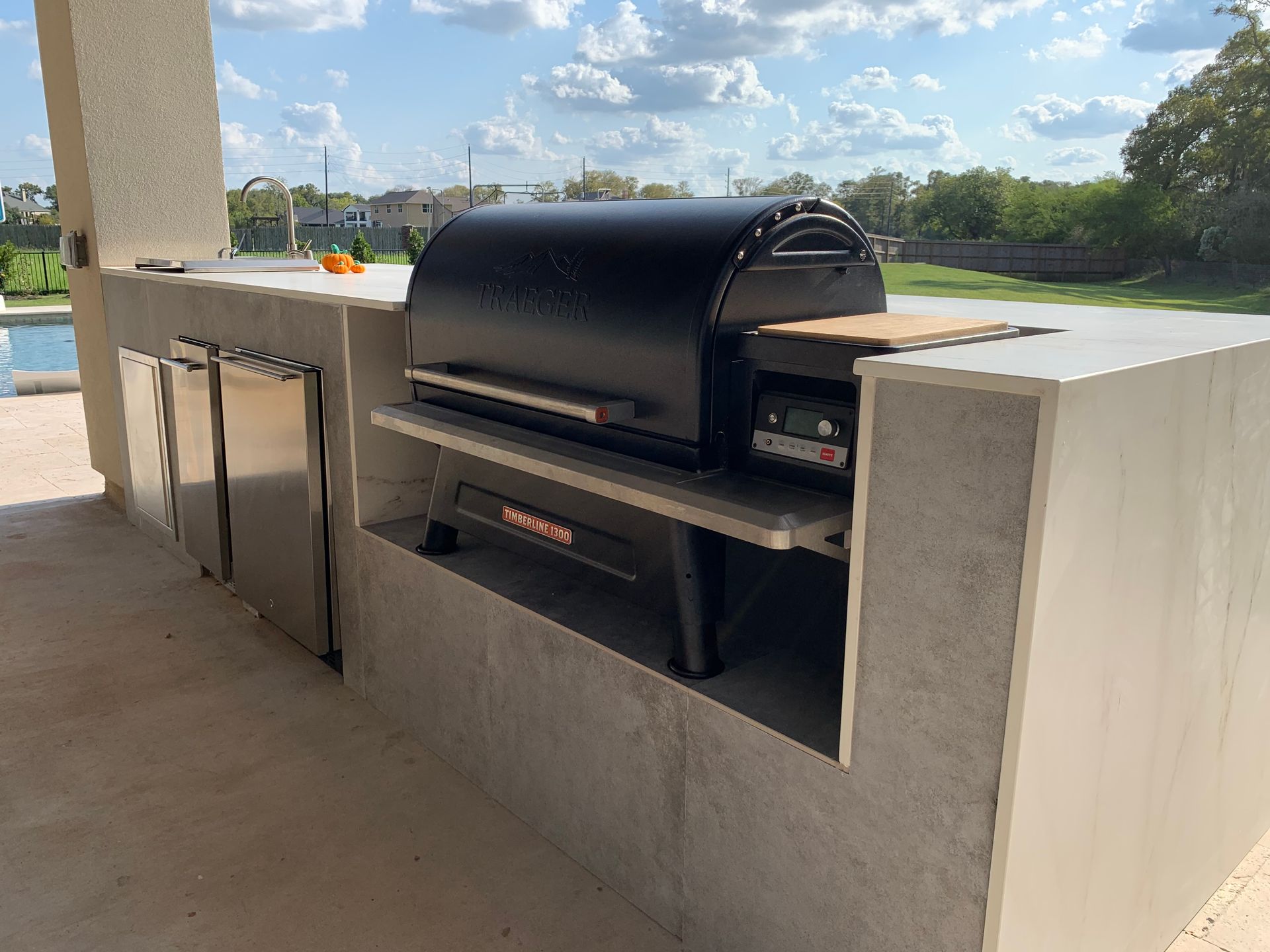 Built-in outdoor kitchen with a black grill, stainless steel fridge, and concrete countertops under a blue sky.