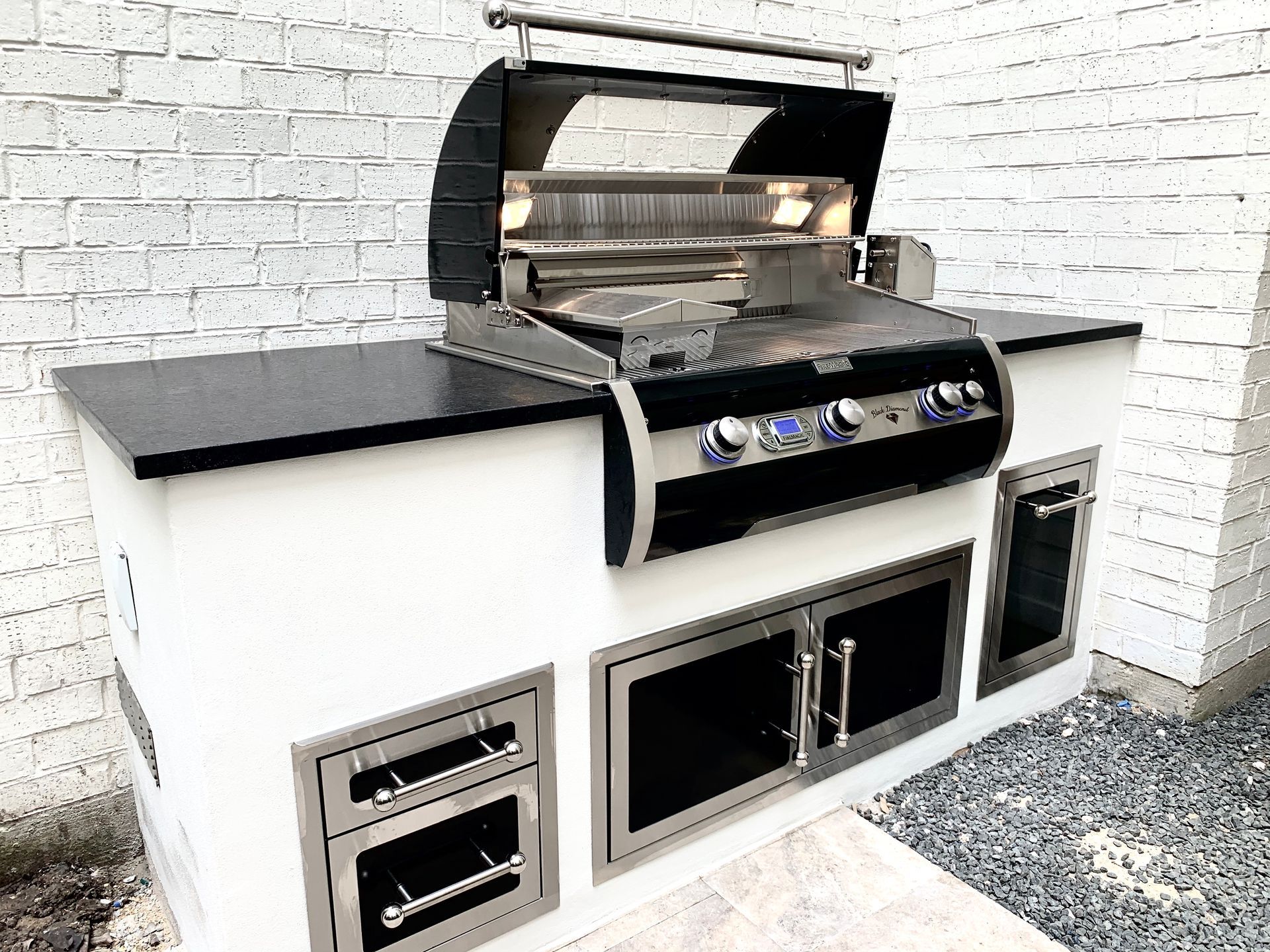 Outdoor kitchen with stainless steel grill, black countertop, white base, and brick wall.