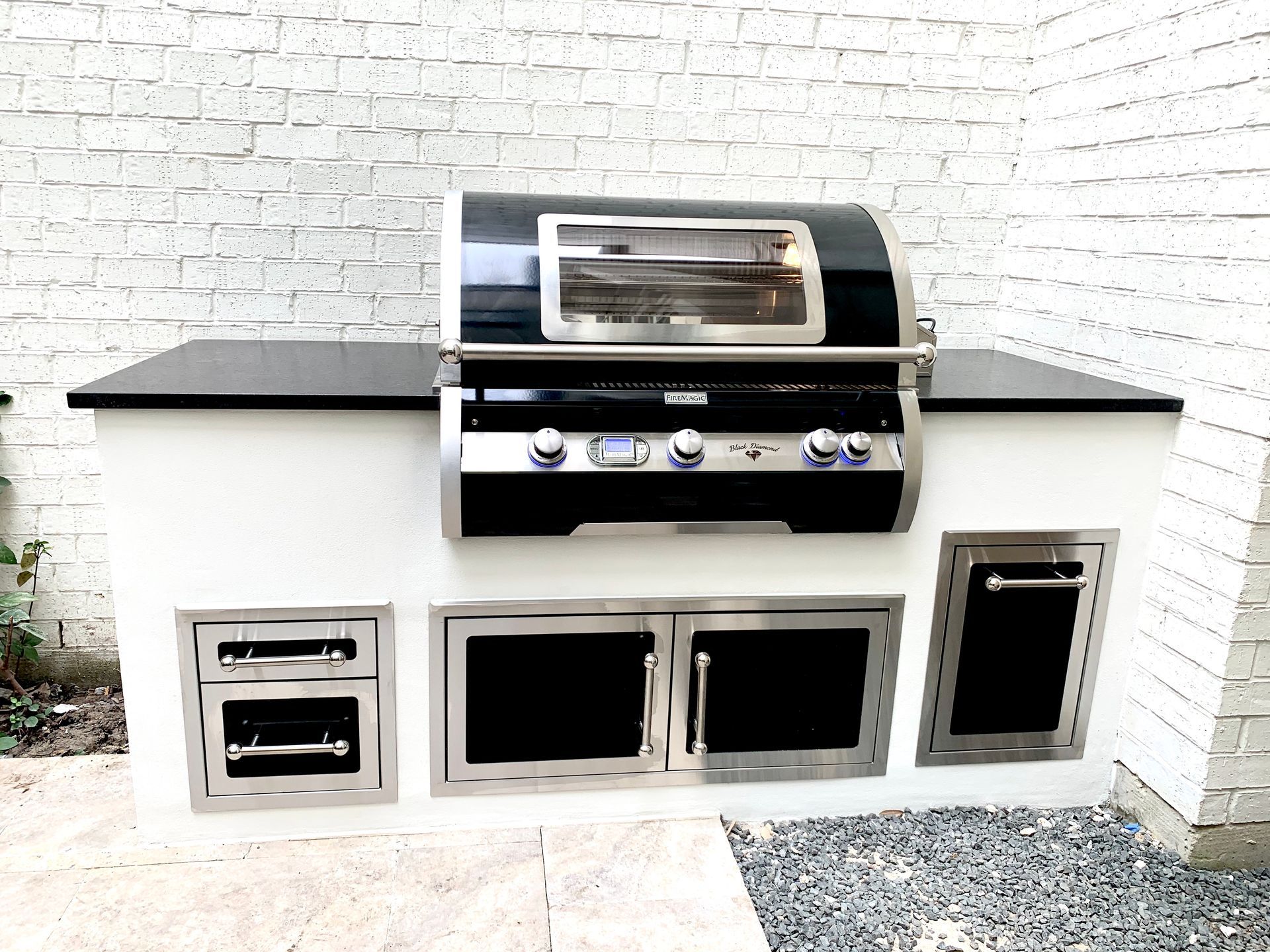 Outdoor kitchen with built-in grill, black countertop, white base, stainless steel doors and drawers, against a brick wall.