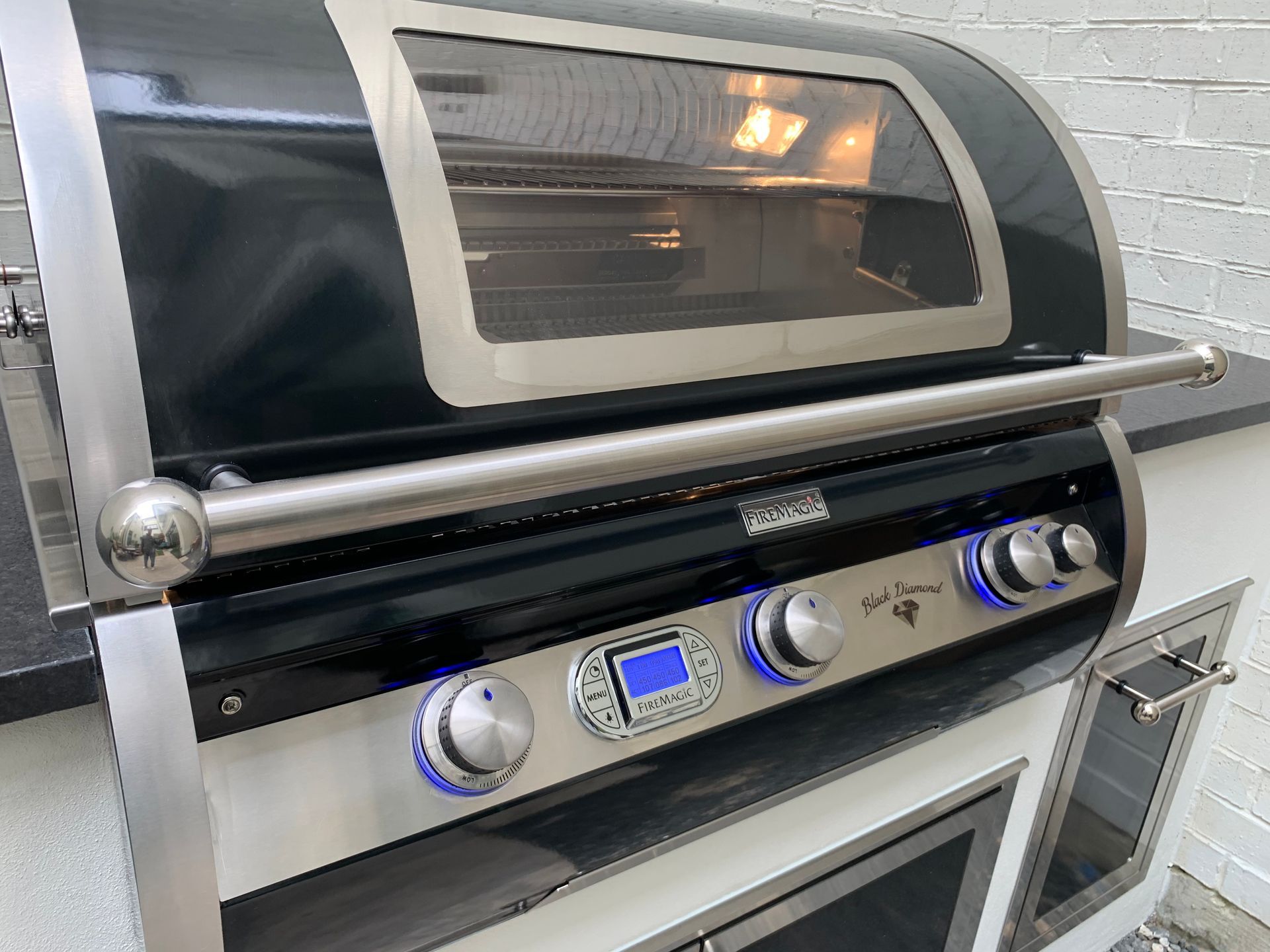 Outdoor grill with oven, stainless steel, black accents, with illuminated dials and oven interior visible.