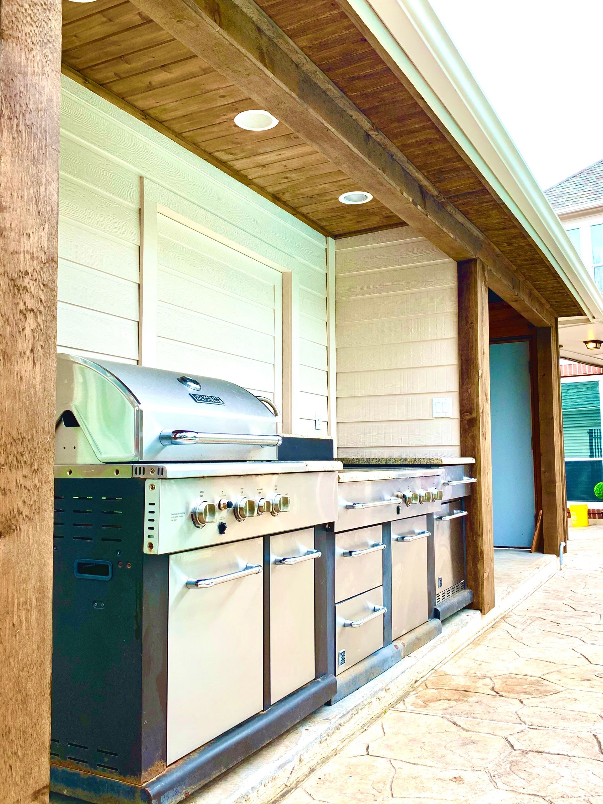 Outdoor kitchen with stainless steel grill and cabinets under a wood-beam covered roof.