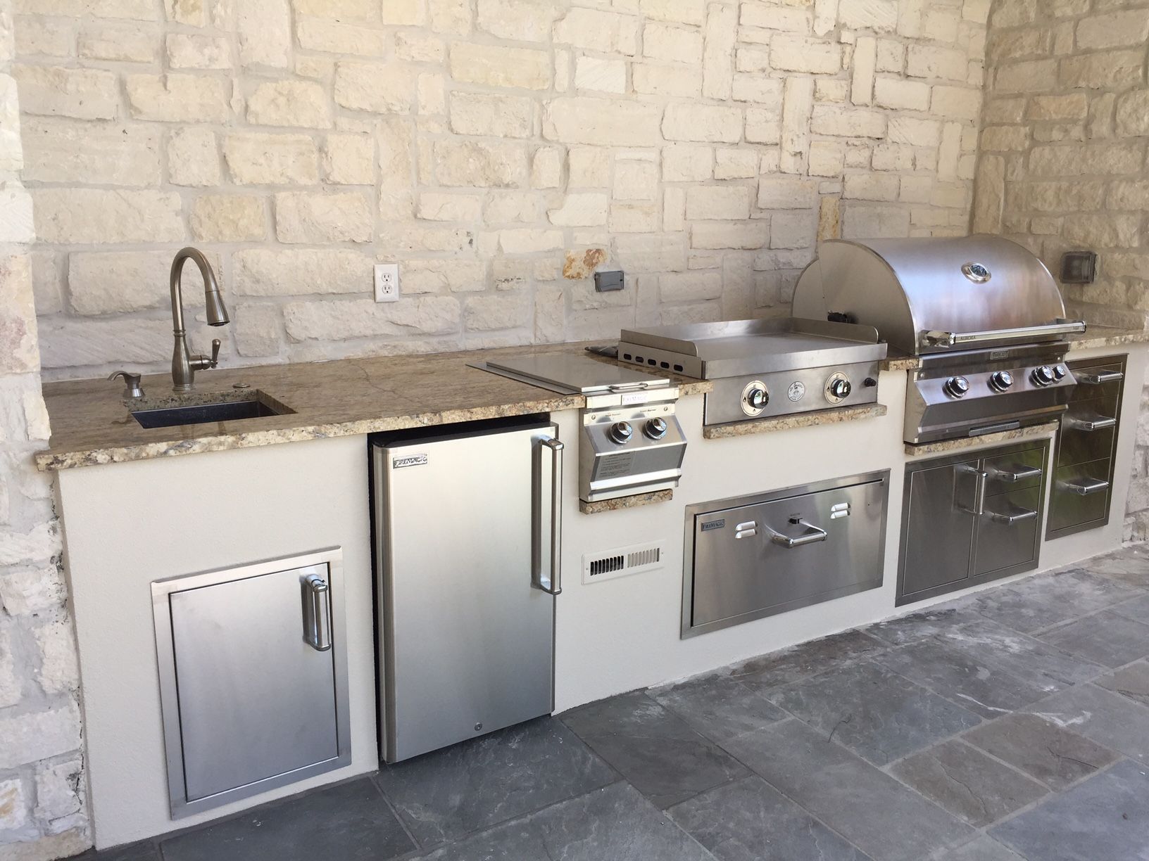 Outdoor kitchen with stainless steel appliances, sink, and stone wall backdrop.