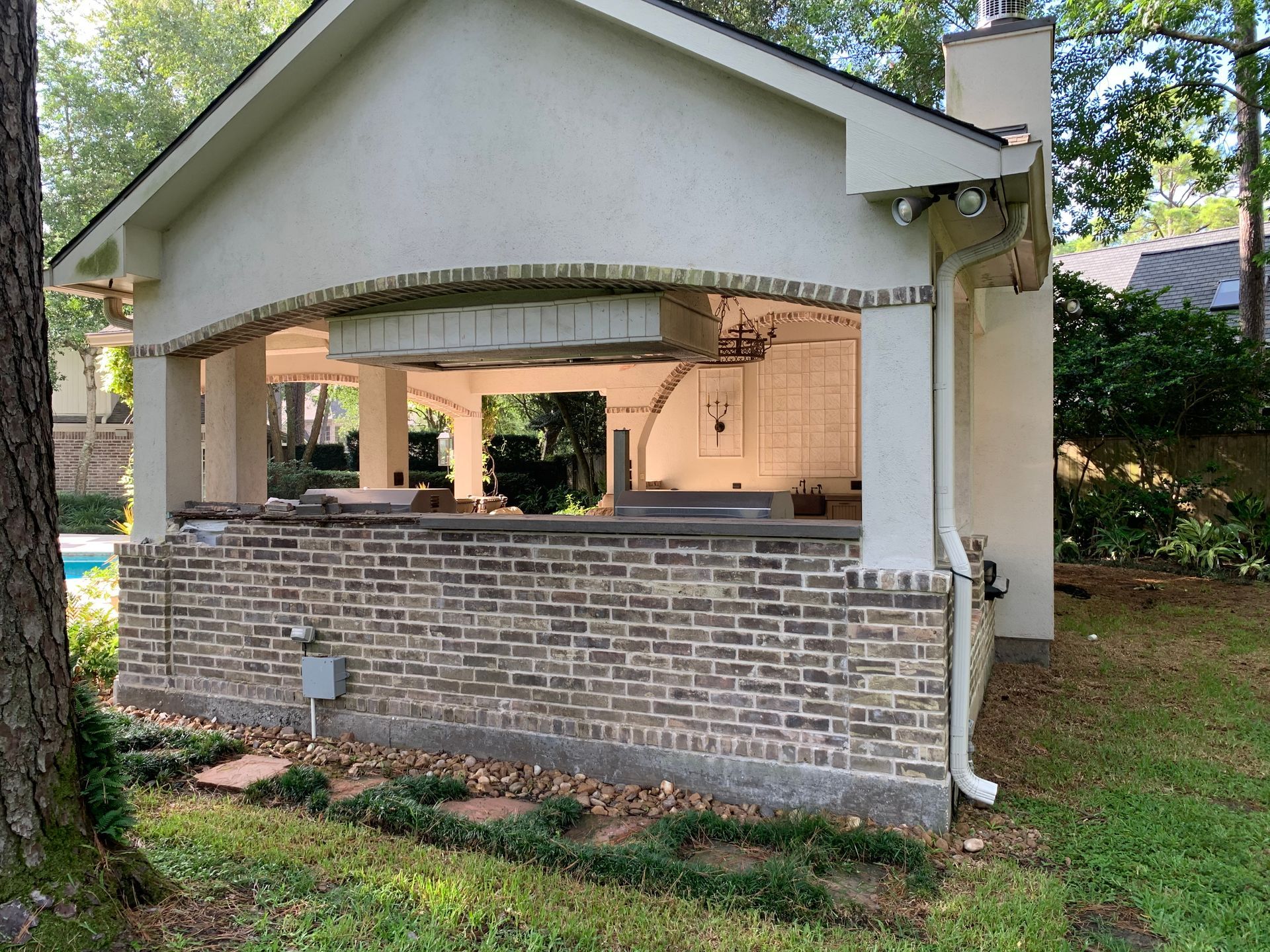 Outdoor kitchen with brick base, arched opening, and stainless steel appliances; lawn and trees in background.