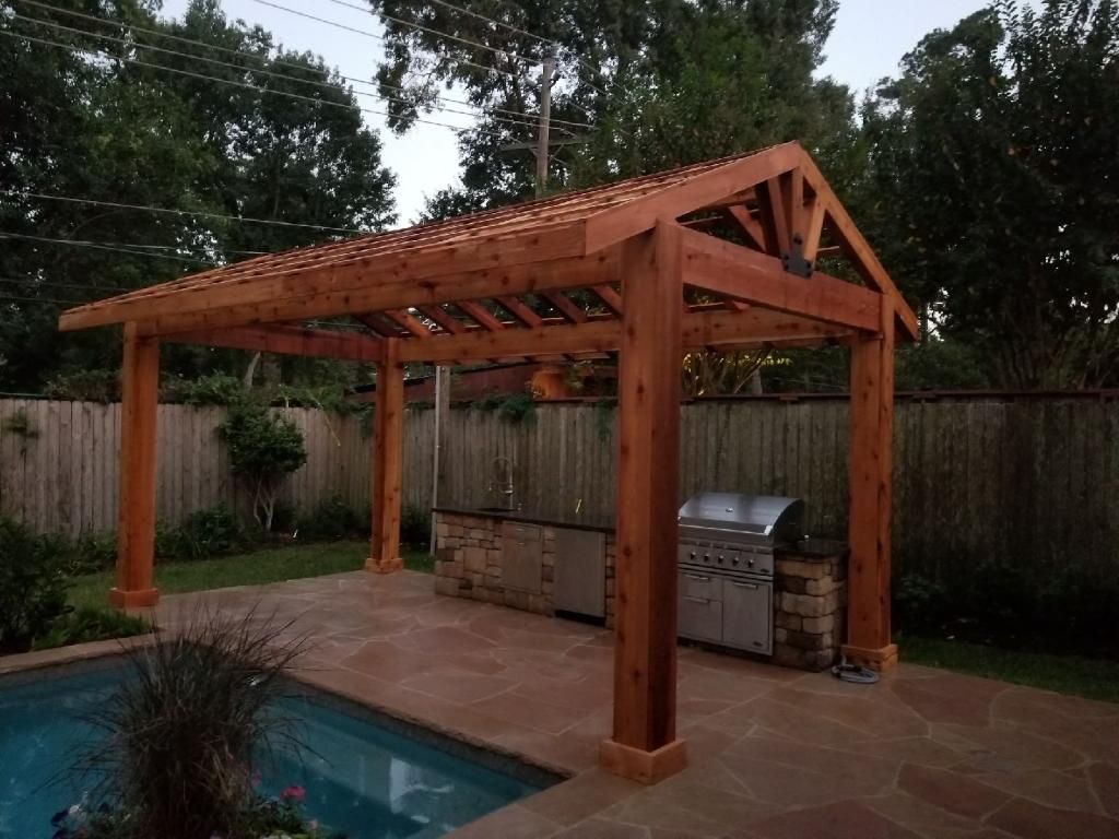 Wooden pergola over outdoor kitchen with pool in the foreground and a wooden fence in the background.