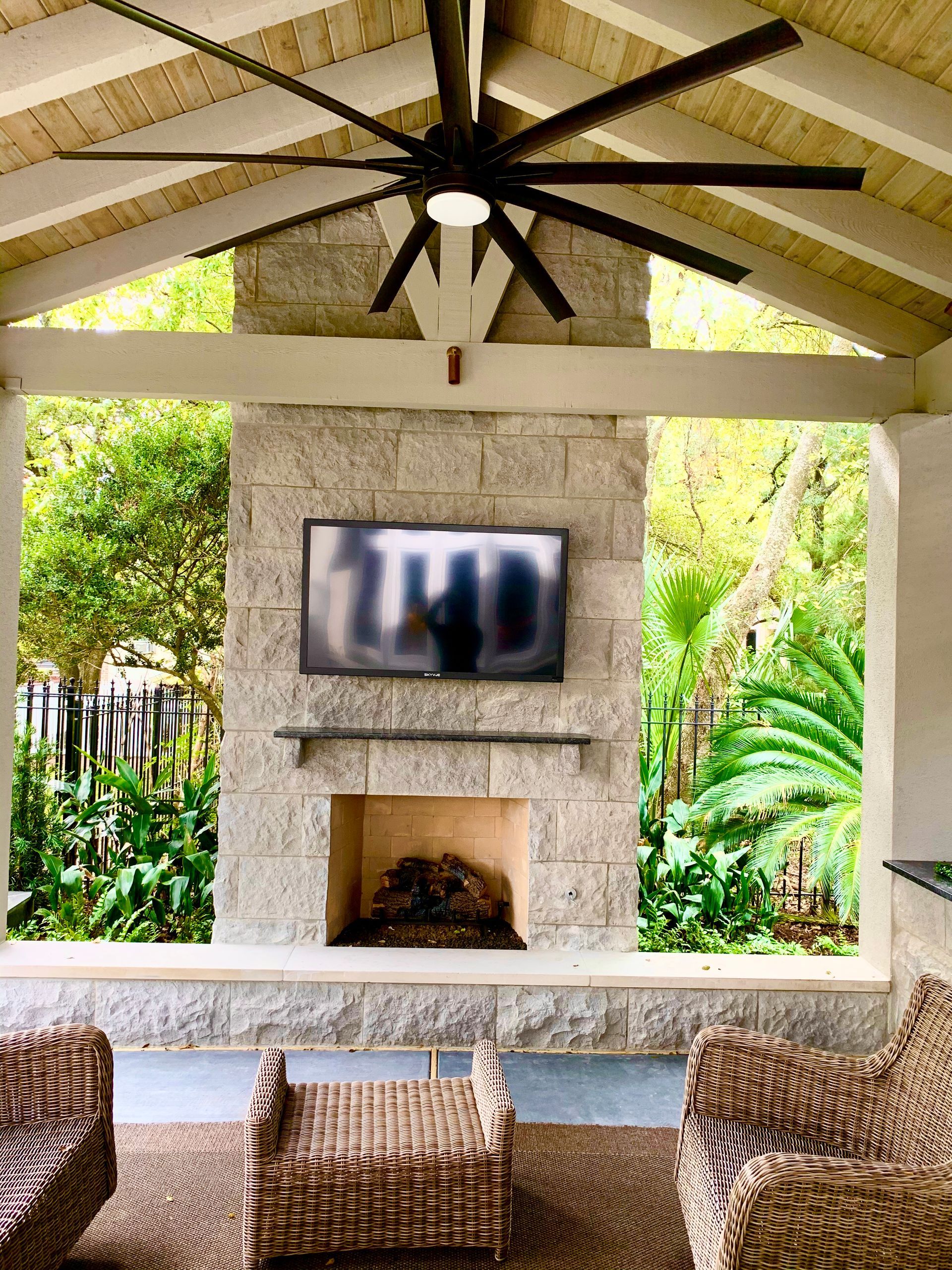 Outdoor patio with stone fireplace, TV, and ceiling fan; wicker furniture sits on a rug.