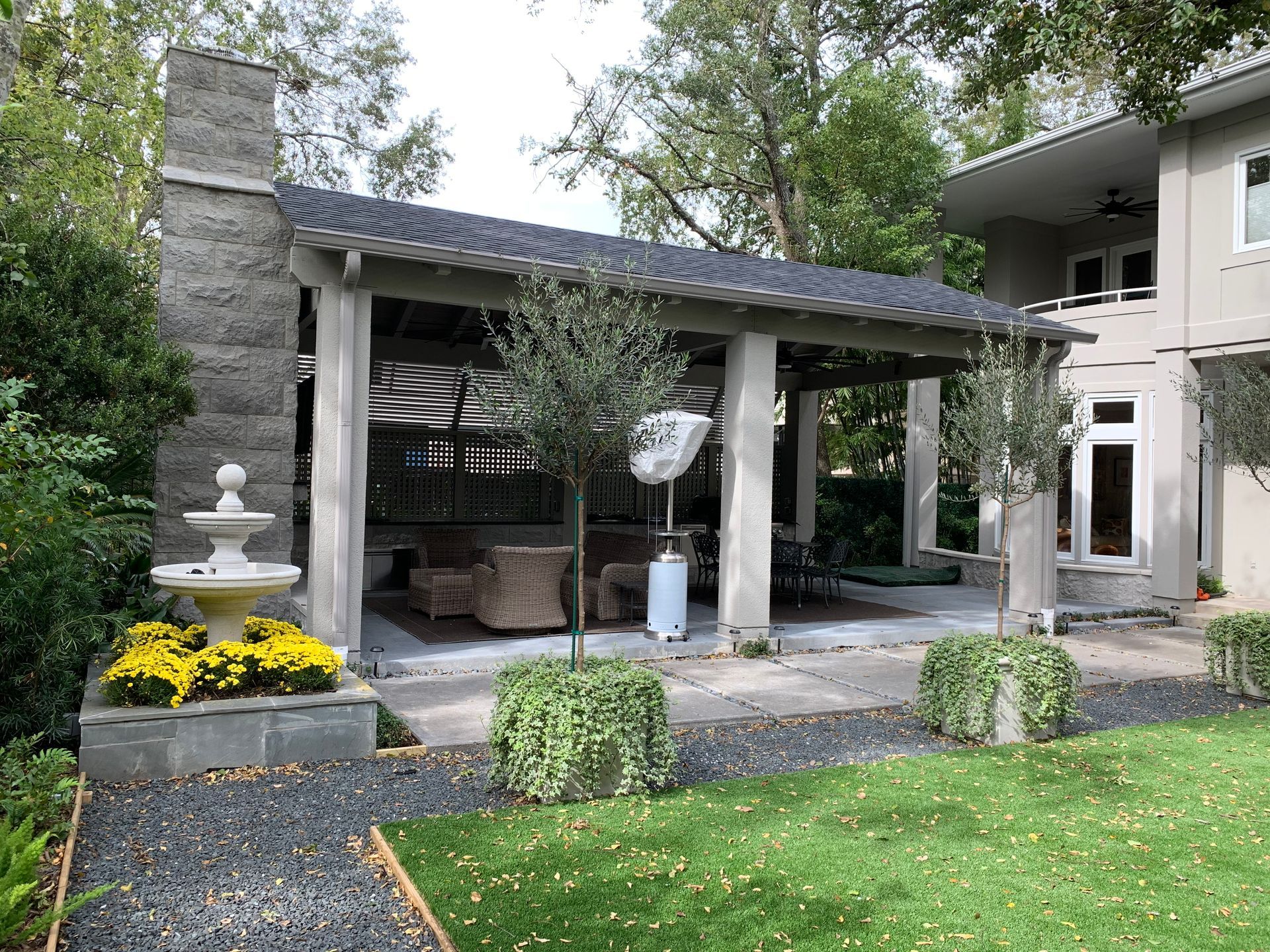 Covered patio with fireplace, fountain, and potted plants, next to a two-story house.