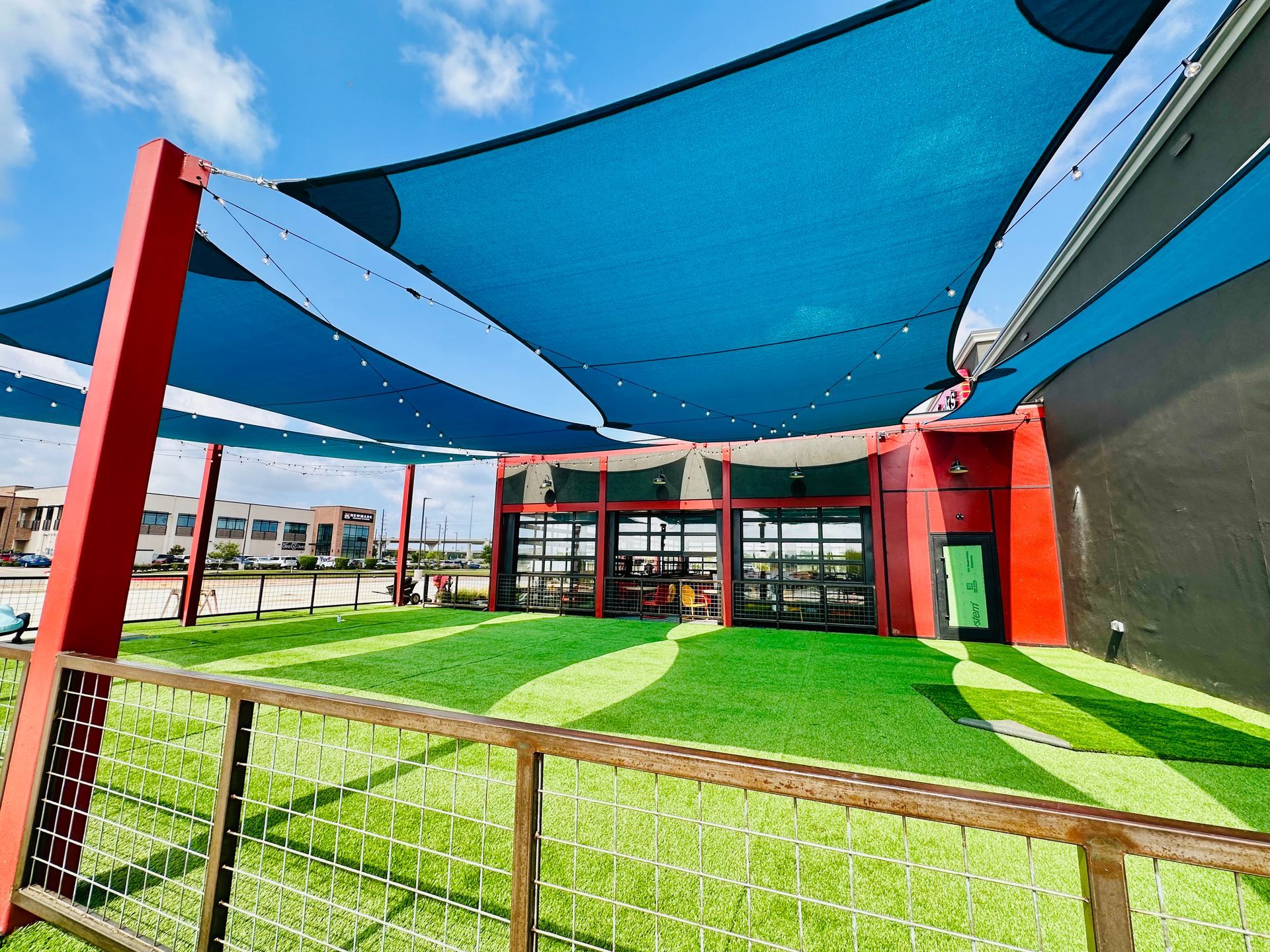 Outdoor dining area with blue shade sails, red supports, glass doors, and turf.