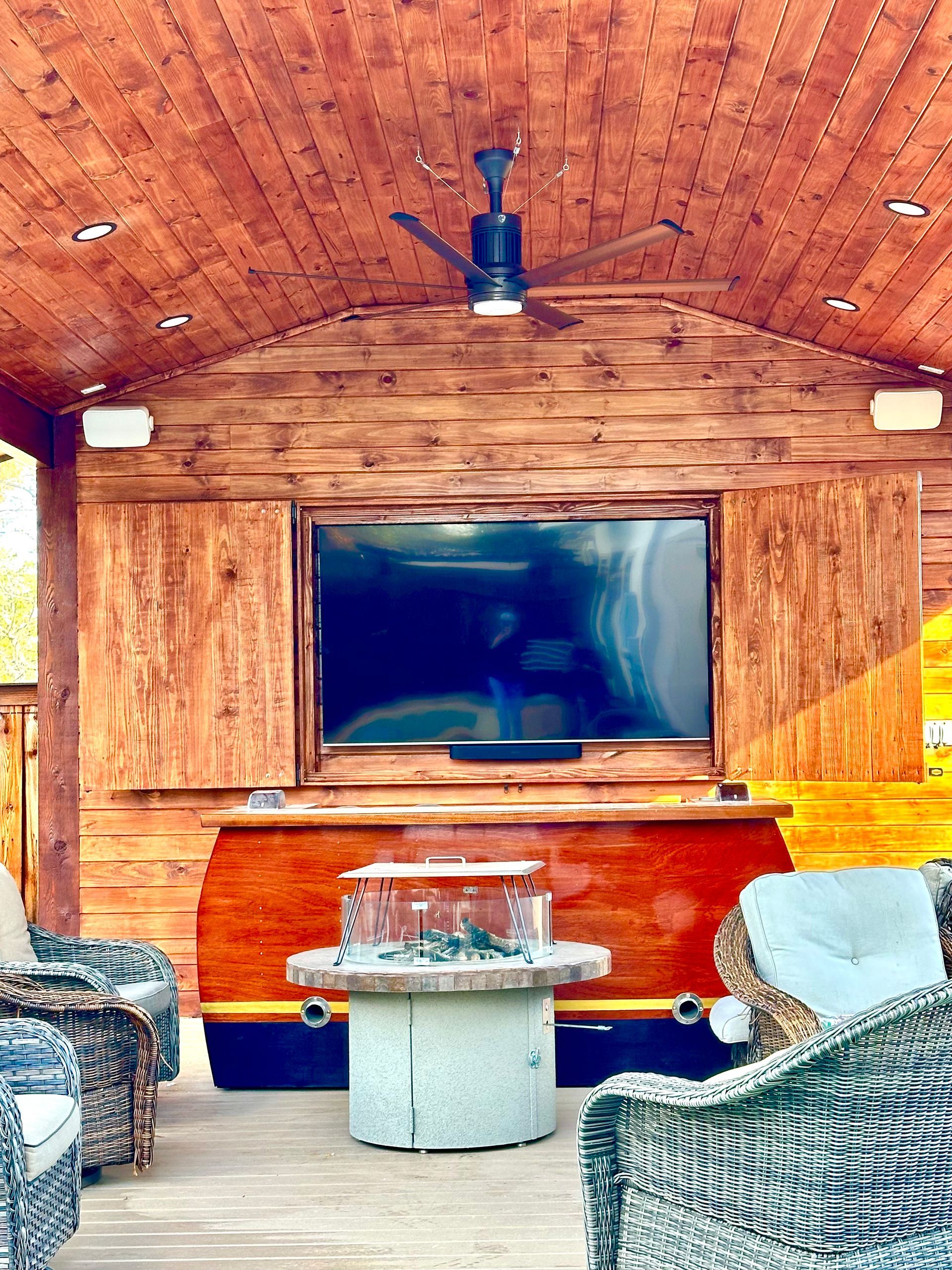 Covered outdoor living space with TV, fireplace, seating, and ceiling fan.