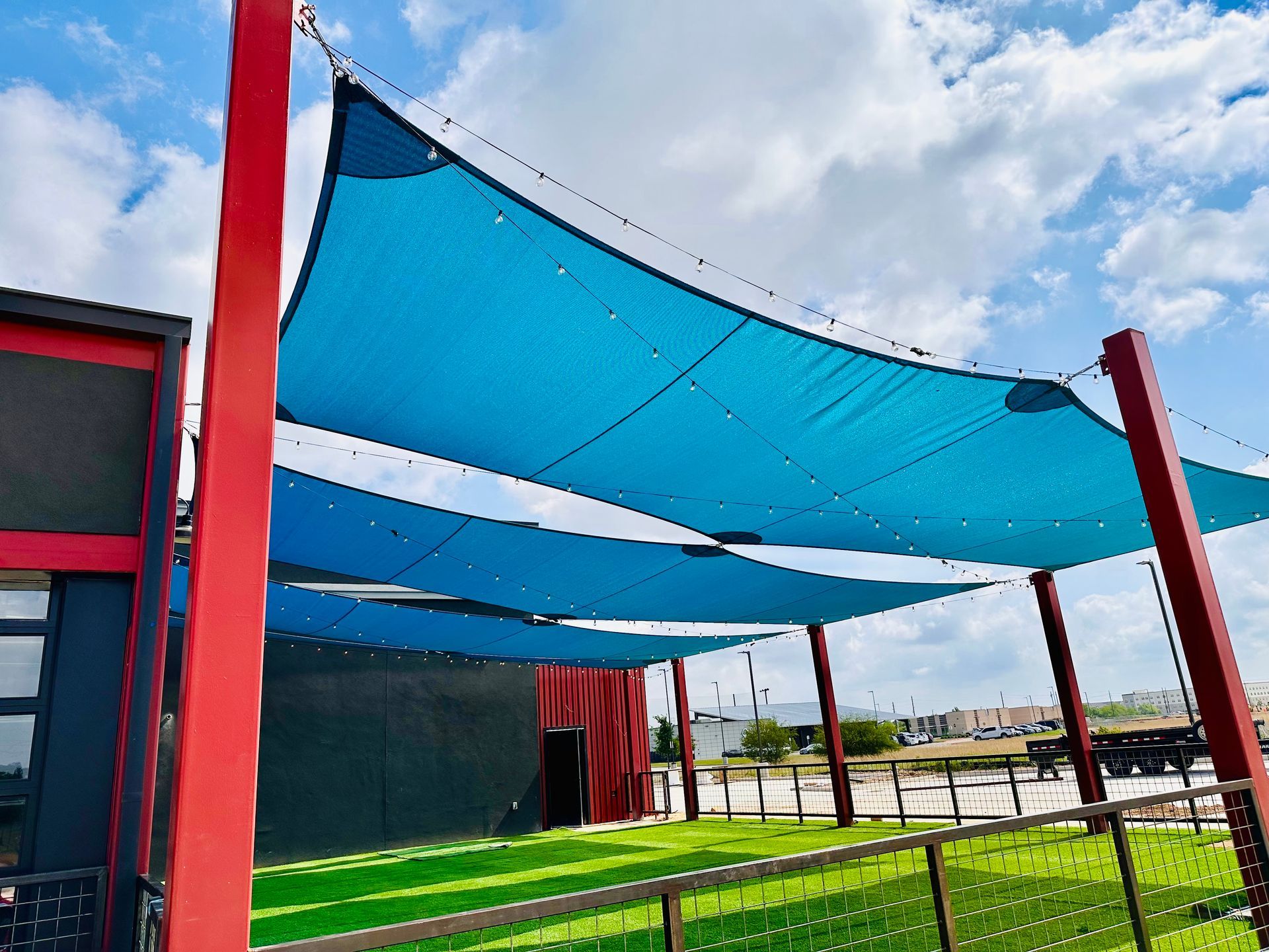 Blue shade sails over a green artificial lawn, supported by red posts.