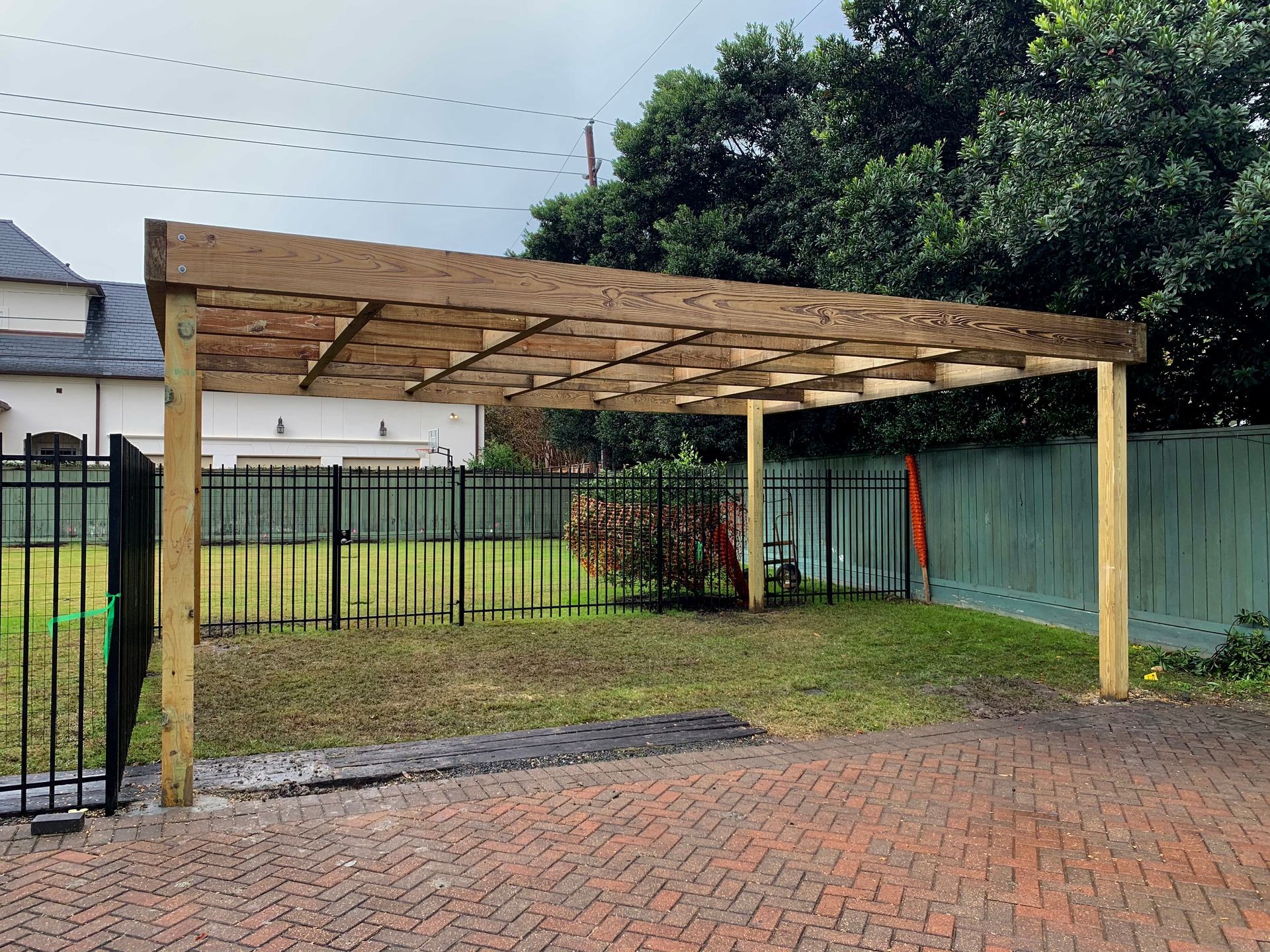 Wooden carport structure in a yard with a brick driveway.