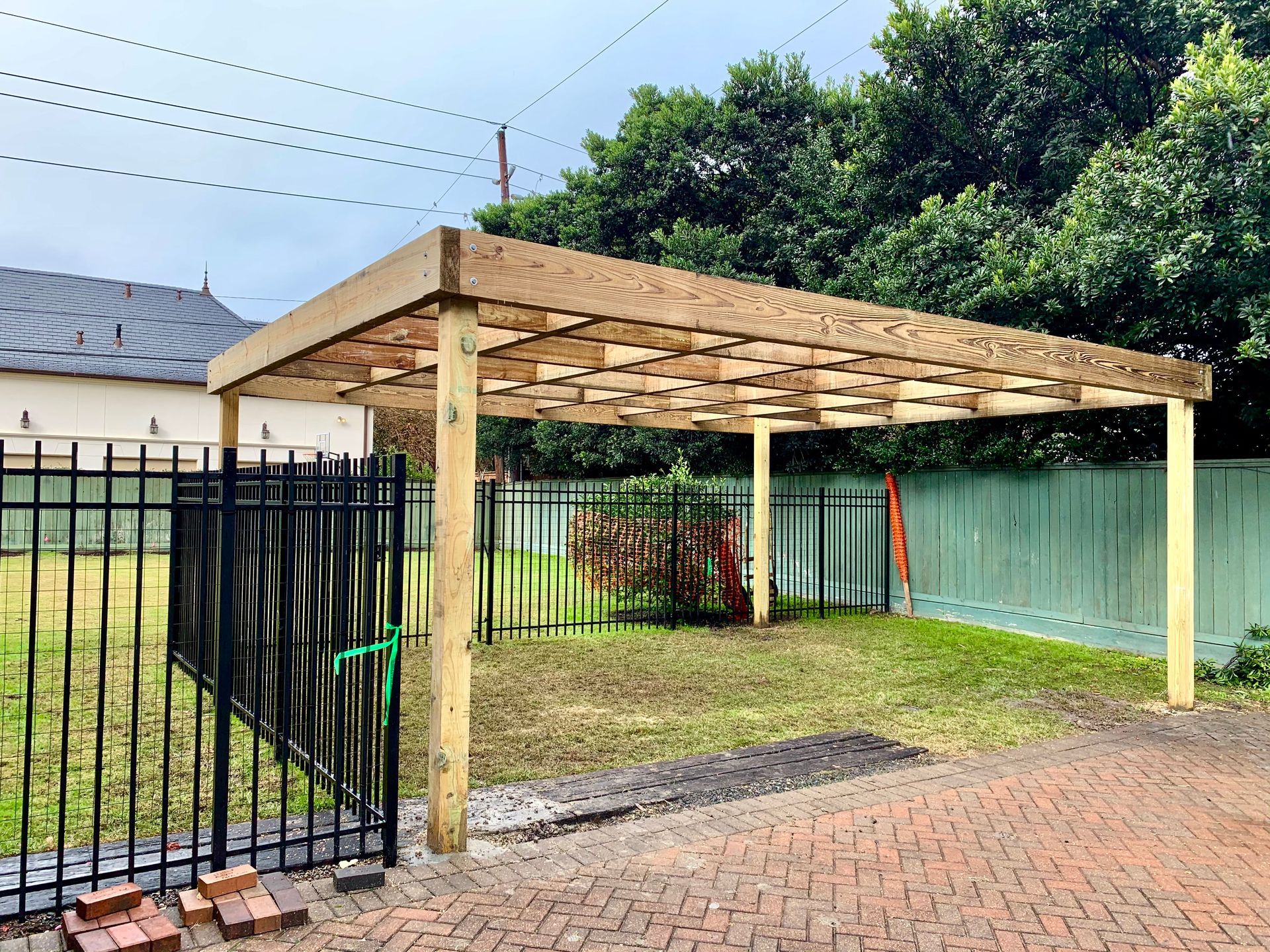 Wooden carport in a yard, with a black fence, green grass, and a blue fence.
