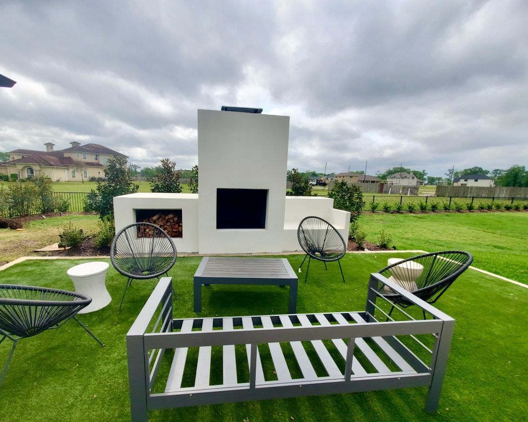 Outdoor seating area with white fireplace, gray furniture, and artificial green grass under a cloudy sky.