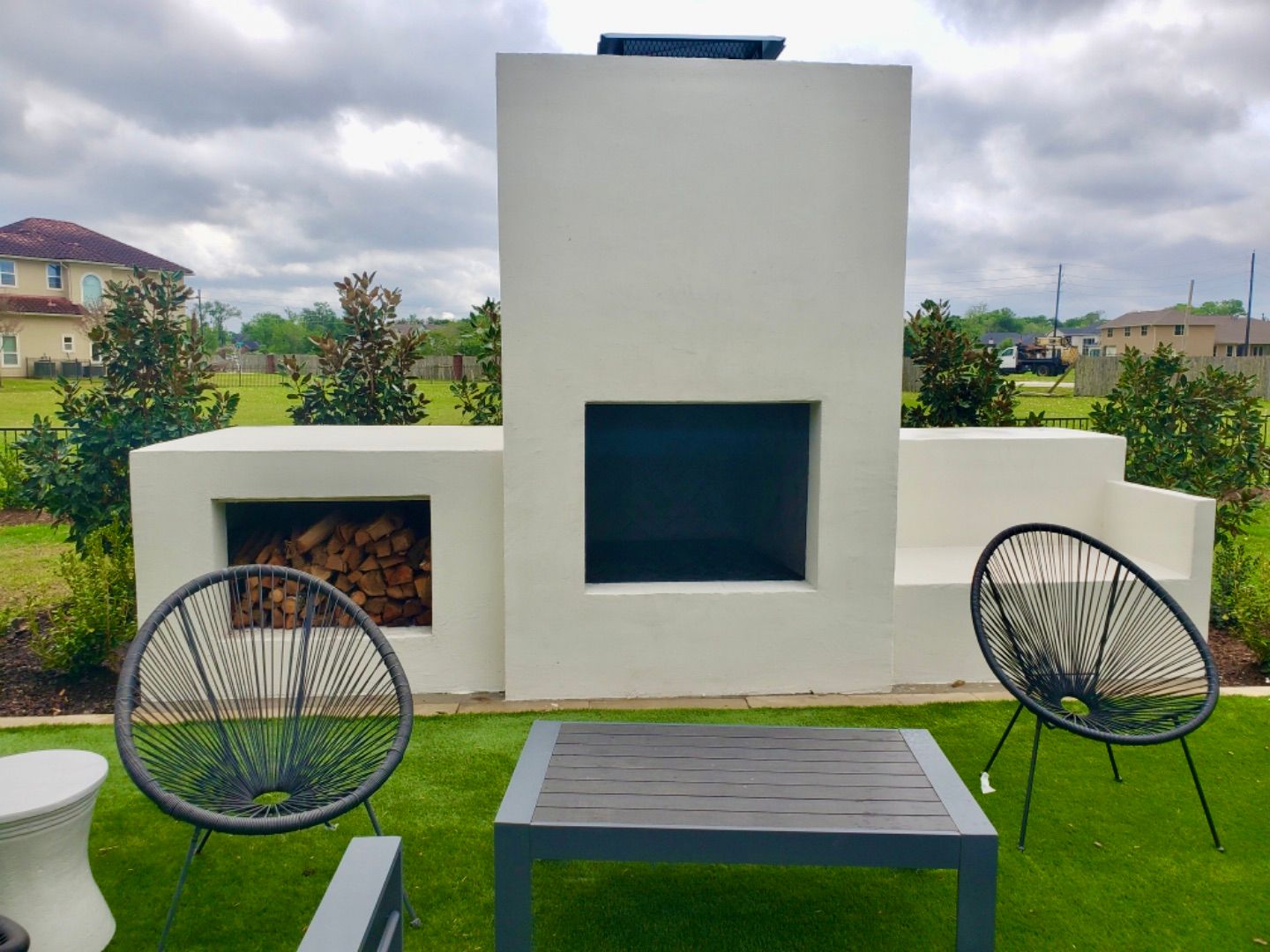 Outdoor fireplace with firewood storage, modern chairs, and table on a lawn.