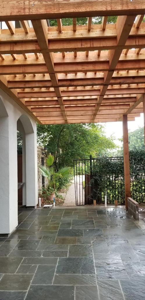 Patio with wooden pergola and stone tile floor leading to a garden gate and foliage.
