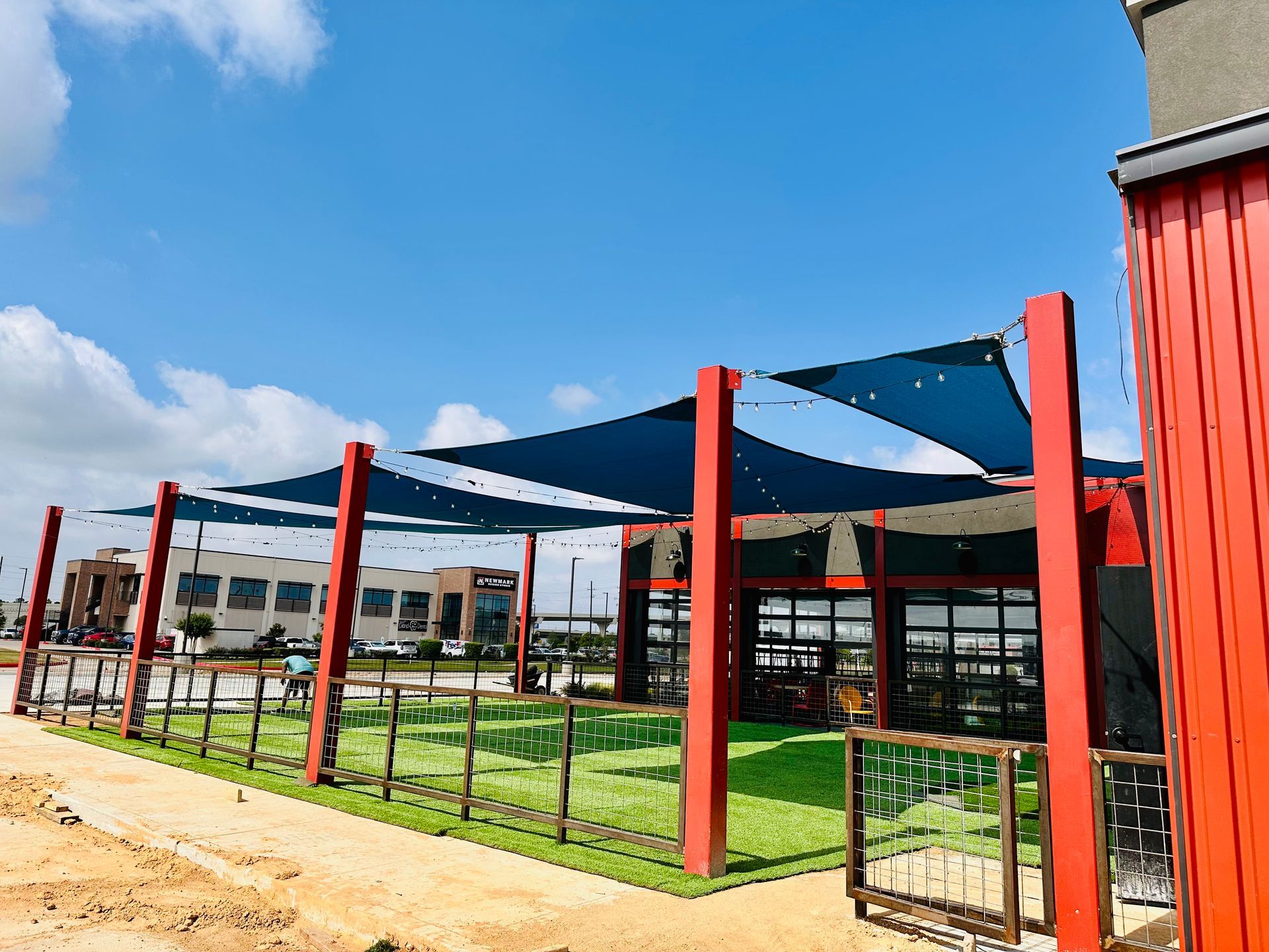 Outdoor seating area with blue shade sails, red pillars, and a green turf lawn.