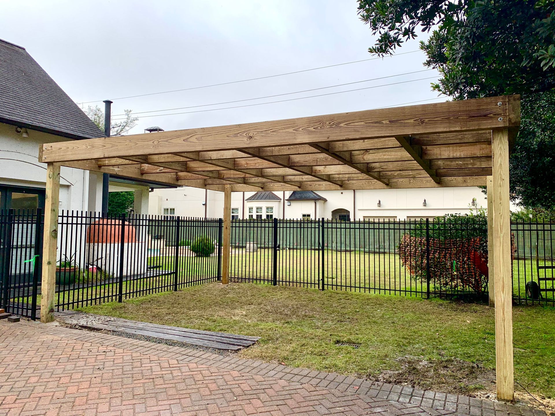 Wooden carport in a yard, supported by posts, over a brick path and grass.