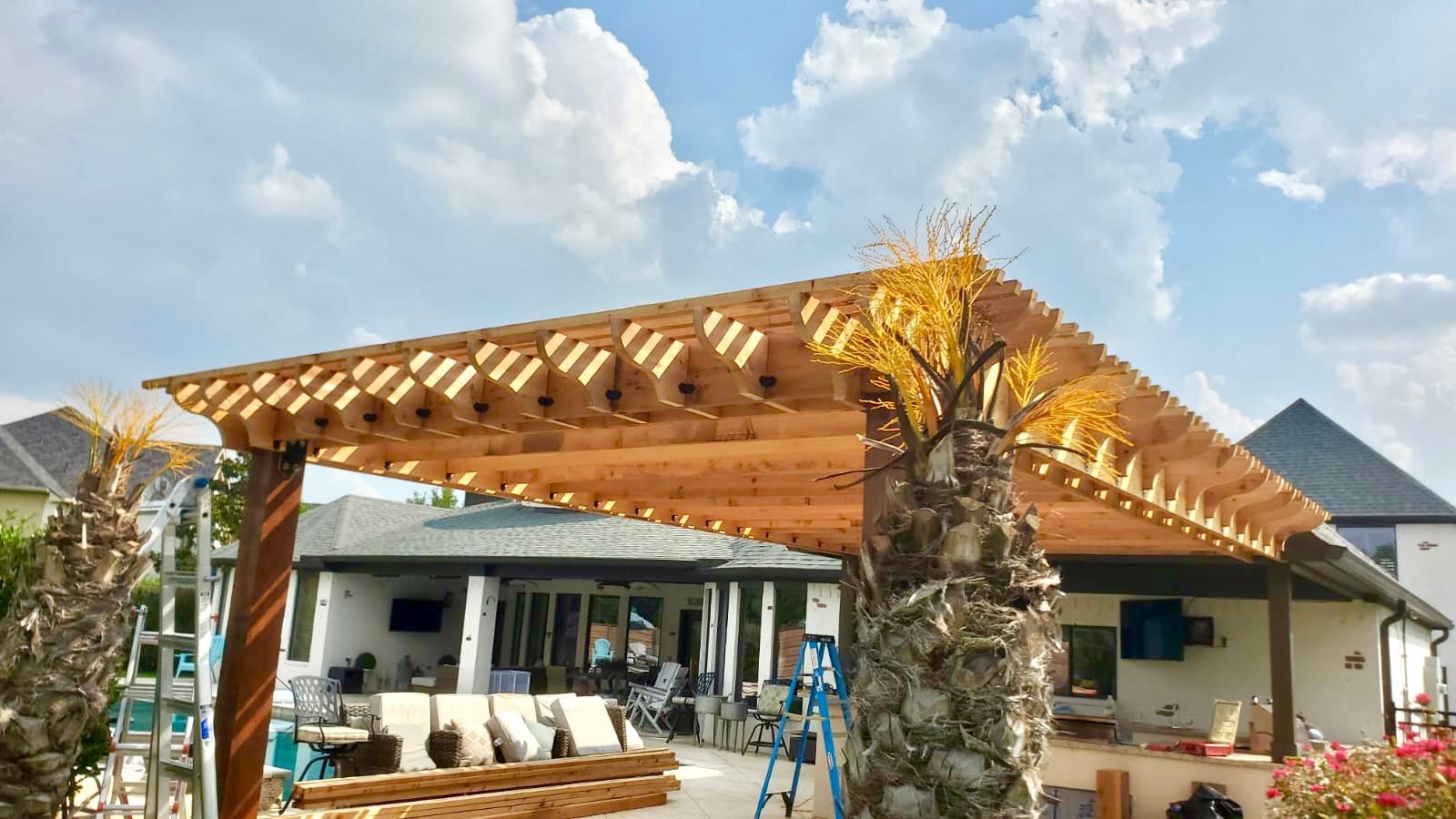 Wooden pergola under construction in a backyard, partially covering a patio; blue sky.