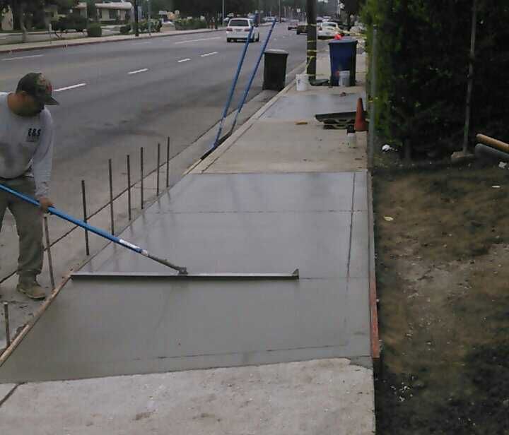 Construction worker smoothing wet concrete sidewalk with a long float, on a street with cars.