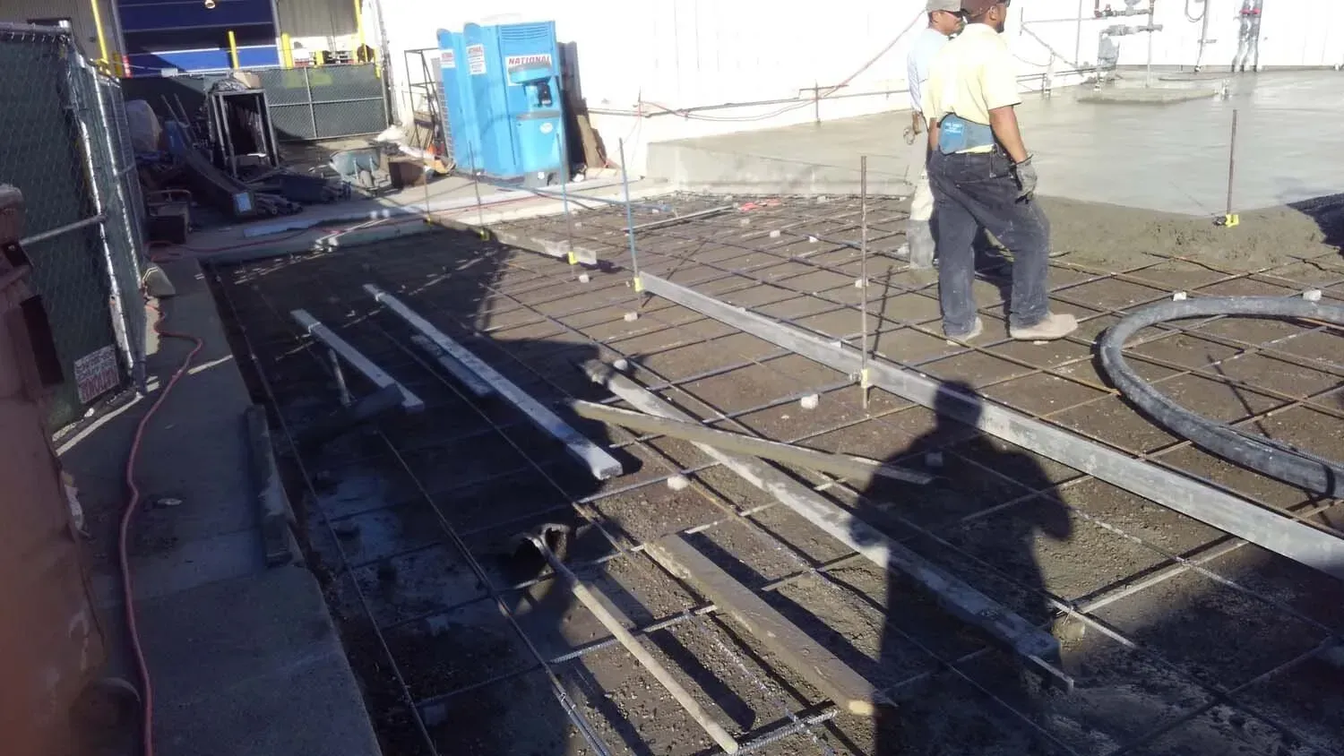 Construction workers pouring concrete on a rooftop, with rebar grid and equipment visible.