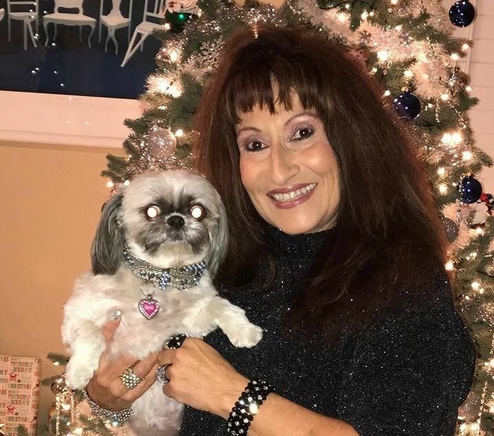 Woman with brown hair smiling, holding a small, white and gray dog in front of a decorated Christmas tree.