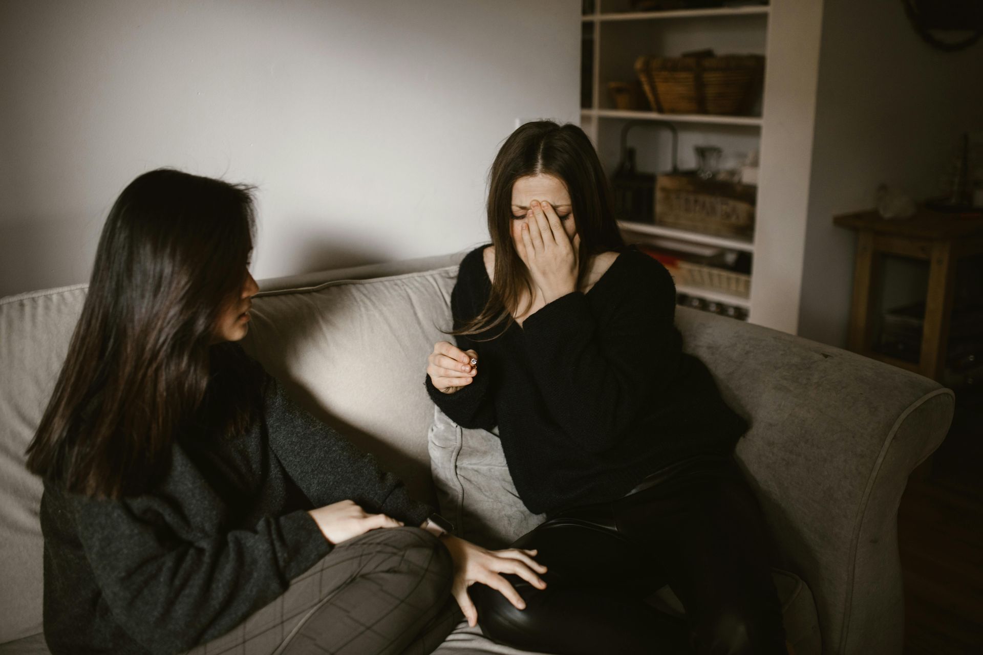 Two women on a couch; one cries, covering her face. The other comforts. Indoors, neutral colors.