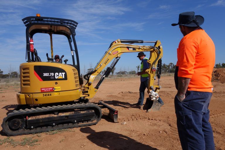 A Man in an Orange Shirt is Standing in Front of a Cat Excavator — Advanced Industry Training In South Townsville, QLD