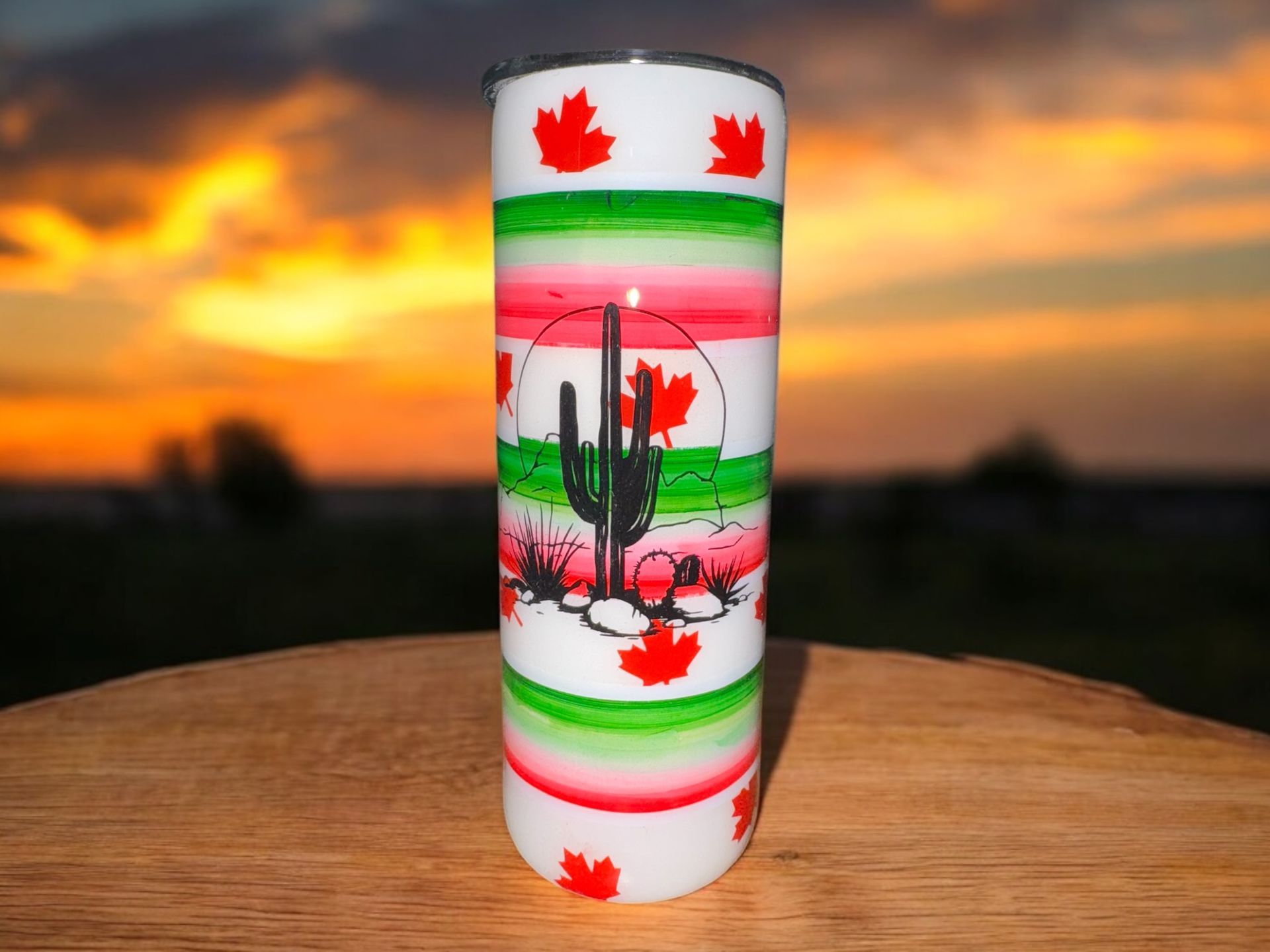 Tumbler with desert cactus design, red and green stripes, maple leaves, on a wooden surface at sunset.