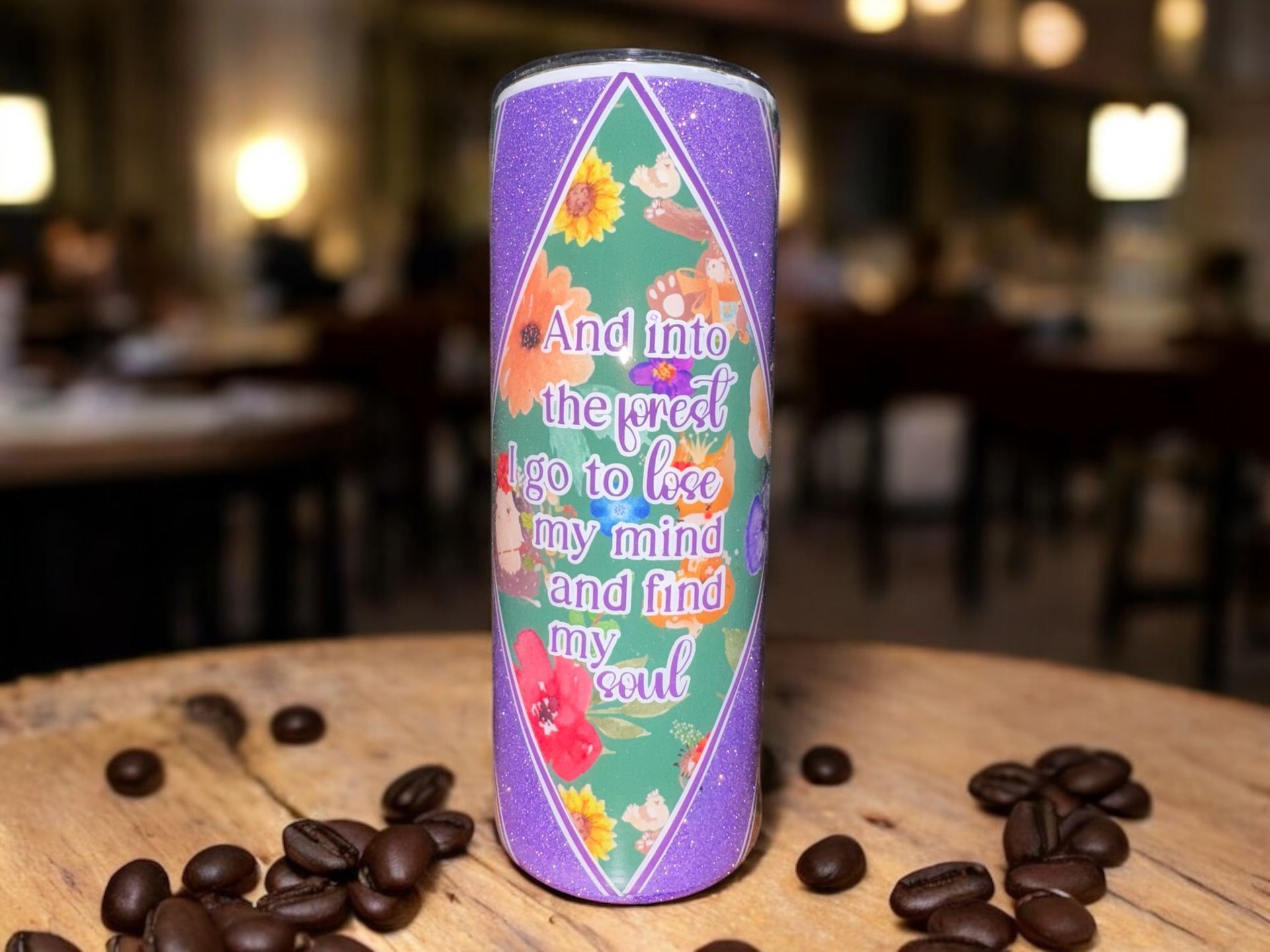 A floral design tumbler with a quote, sitting on a wooden surface with coffee beans scattered around.