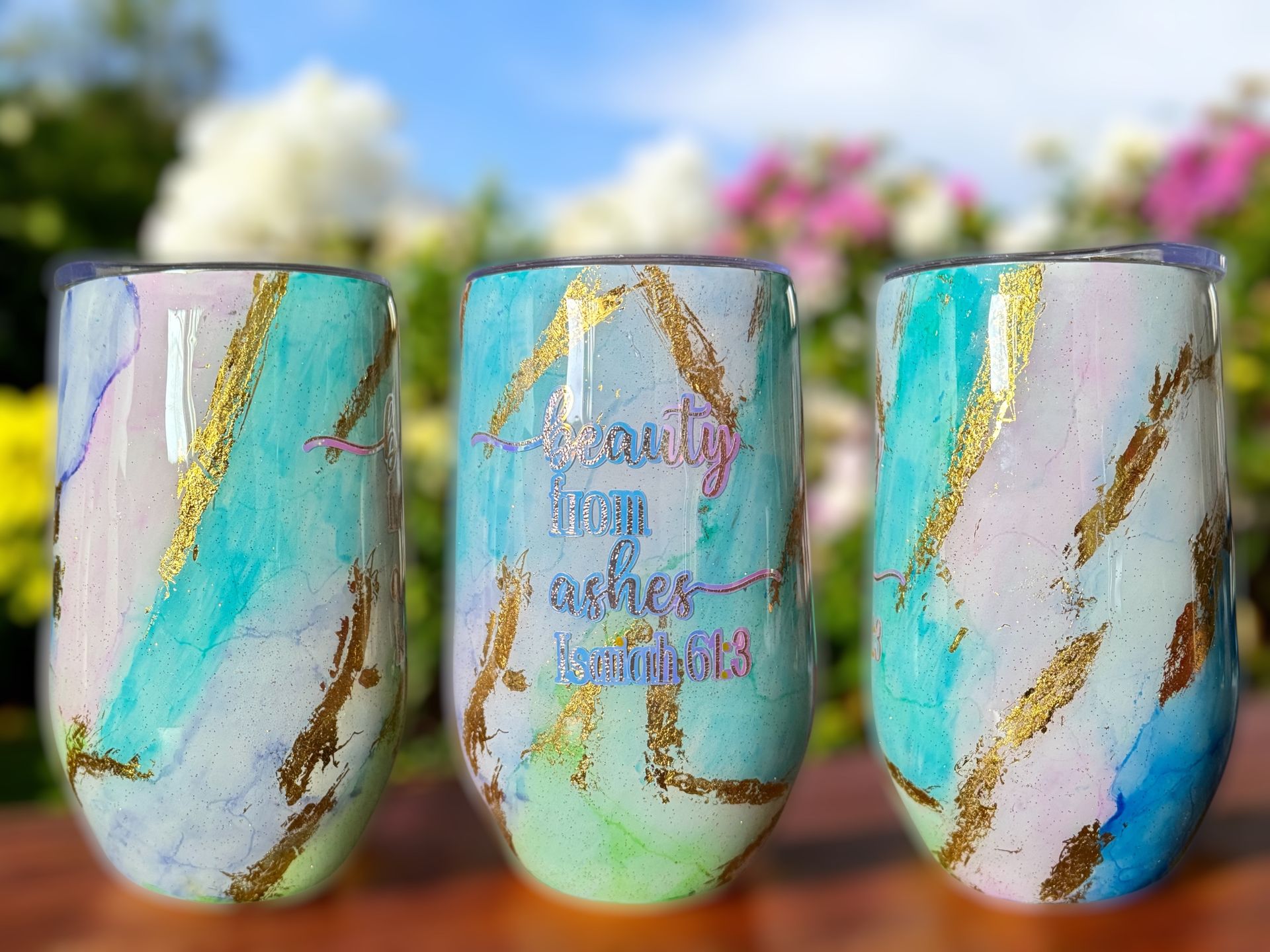 Three stemless wine tumblers with watercolor-like swirls of blue, green, and gold, with gold accents. The center tumbler has the inscription 