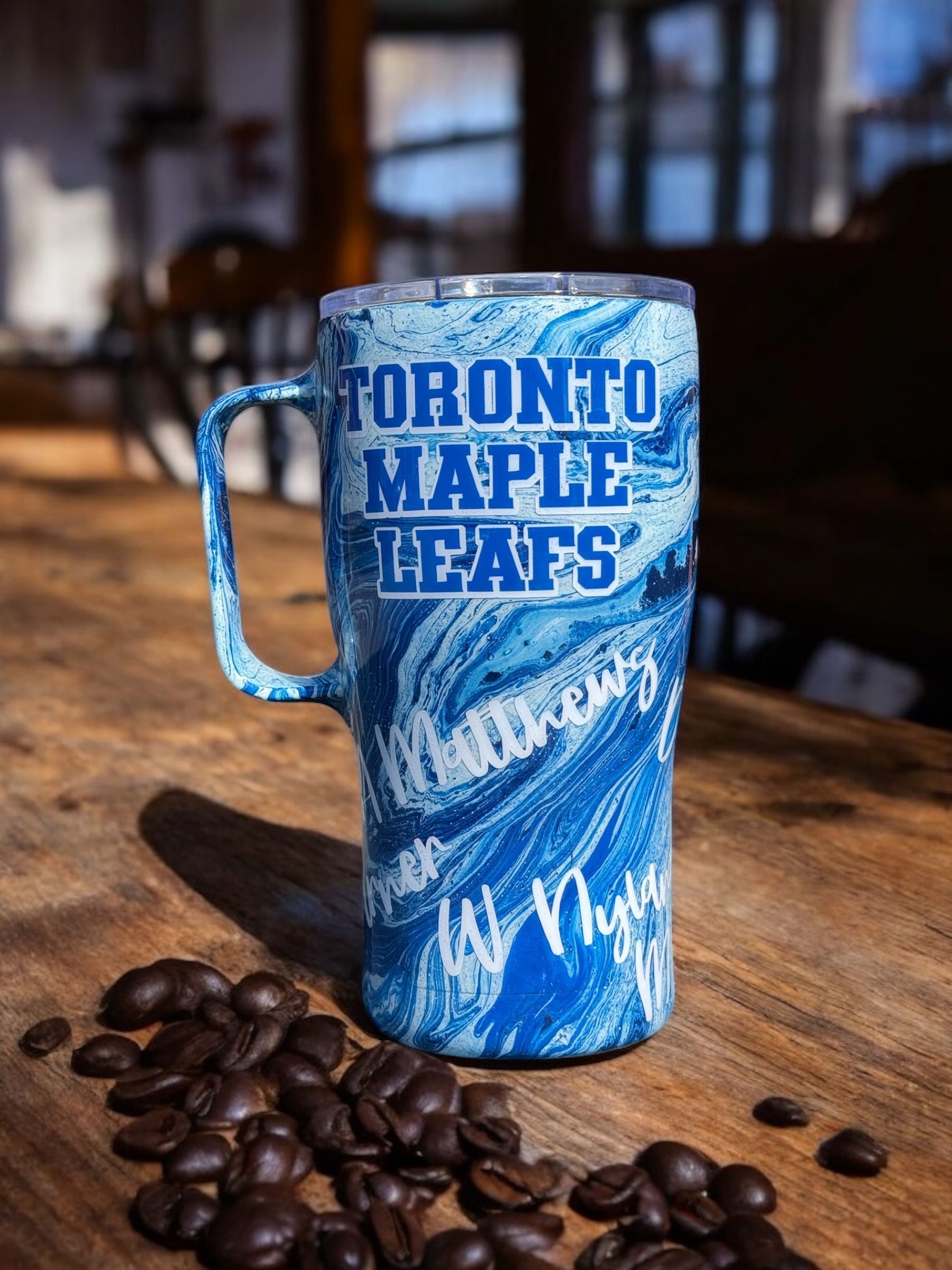 Toronto Maple Leafs tumbler with blue and white swirl design, handle, on wooden table with coffee beans.