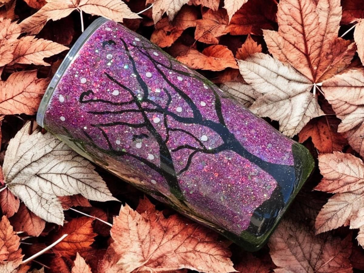 Glittery tumbler with black tree design, set among colorful autumn leaves.