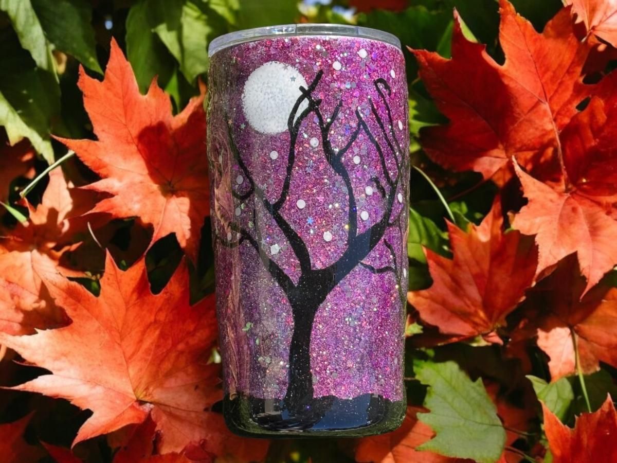 Glittery purple tumbler with a black tree design, surrounded by brown fall leaves.
