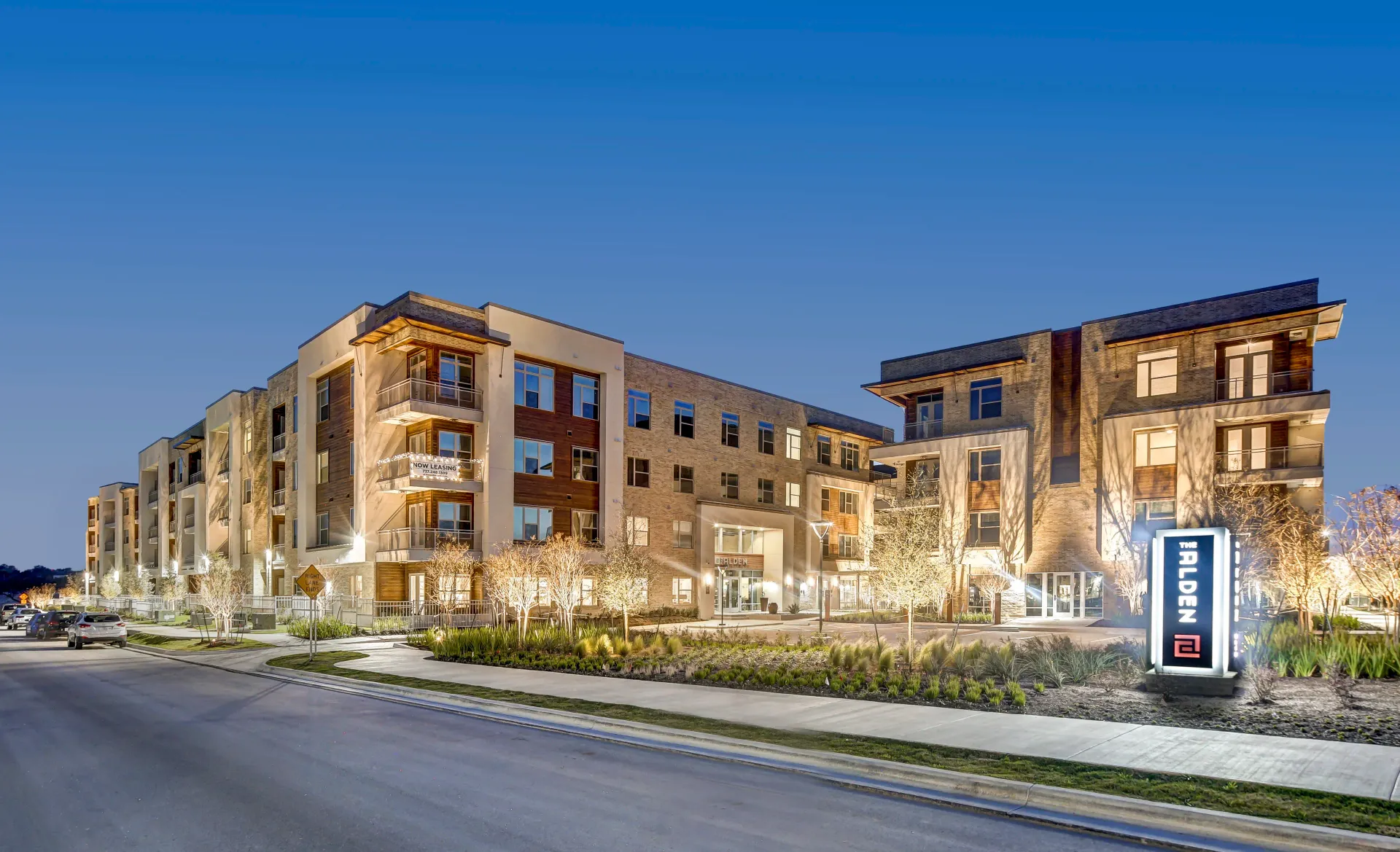 Multi-story apartment building at dusk with exterior lights illuminating facade, landscaping, and signage.