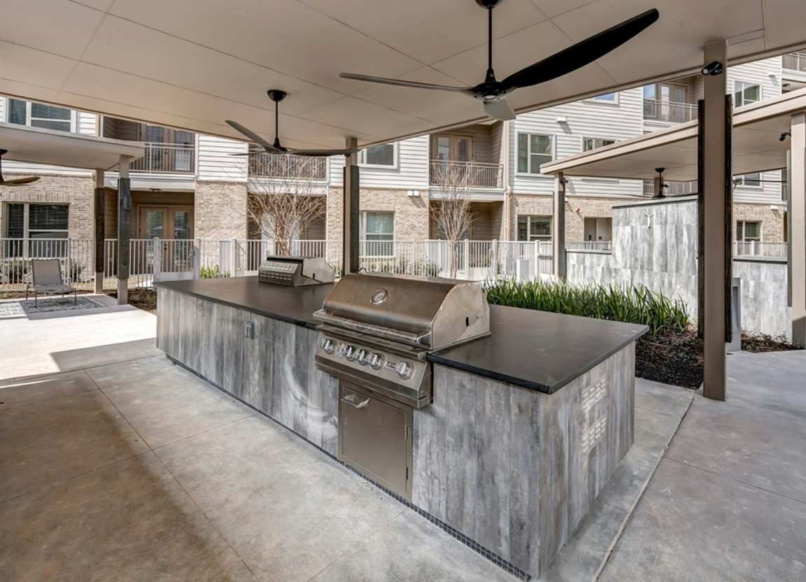 Outdoor grilling area with a built-in grill and countertop under a covered patio. Buildings are visible in the background.