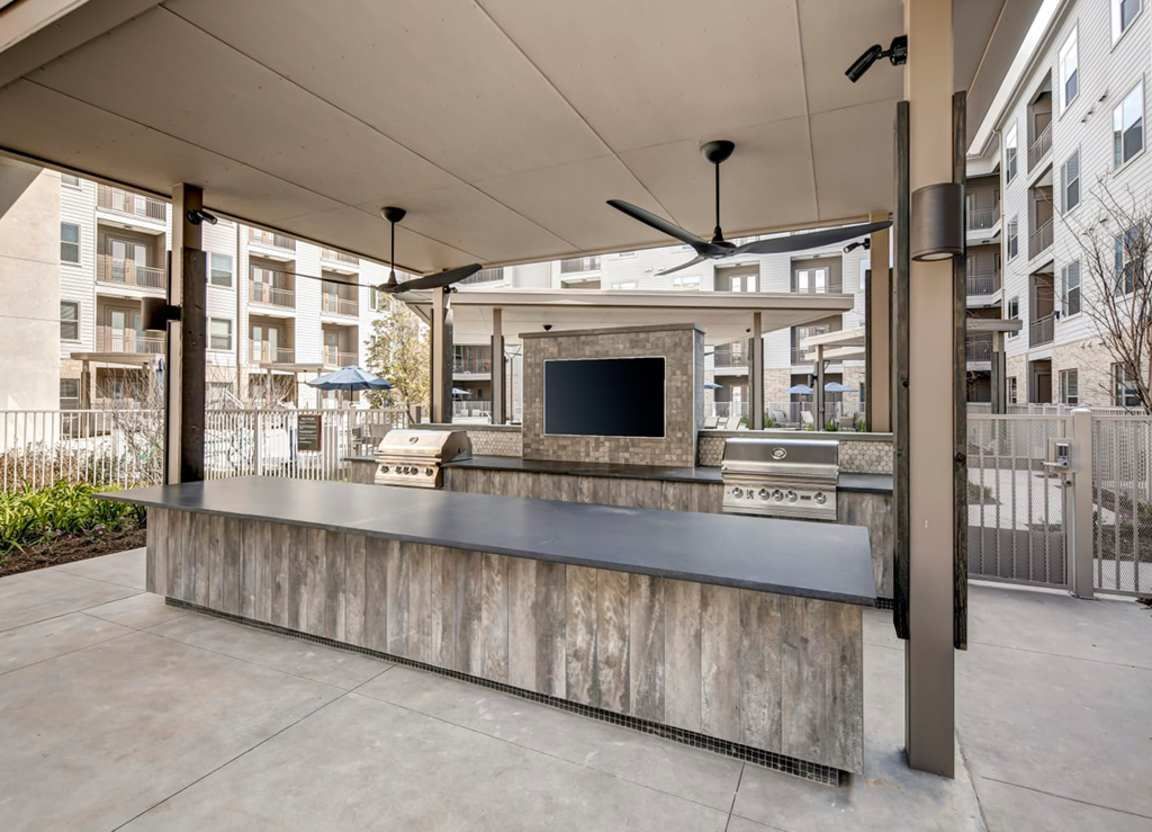 Outdoor grilling area with a long countertop, grills, a TV, and ceiling fans under a covered patio.